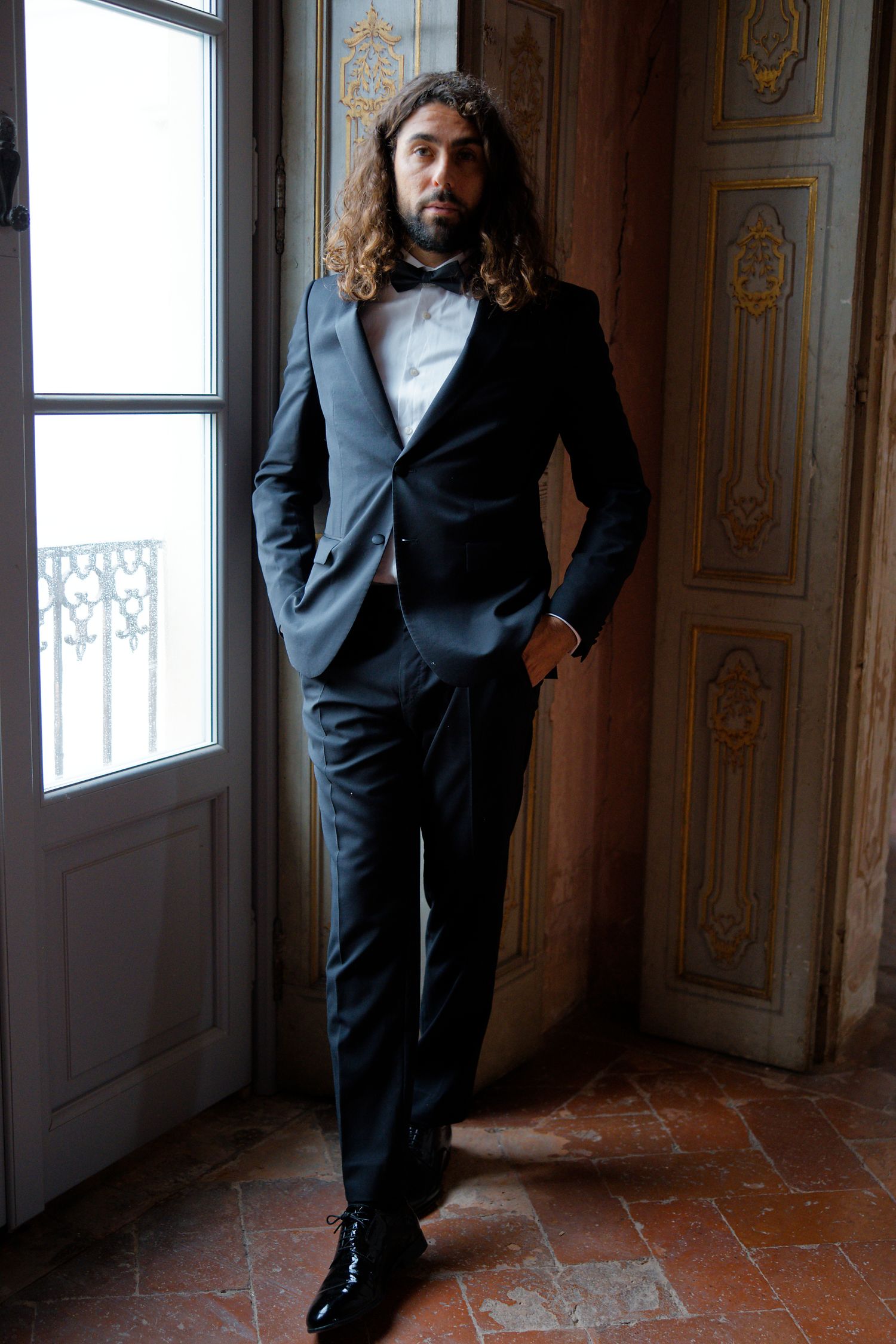 A groom in a black tuxedo poses by a window in an elegant historic room with ornate details.
