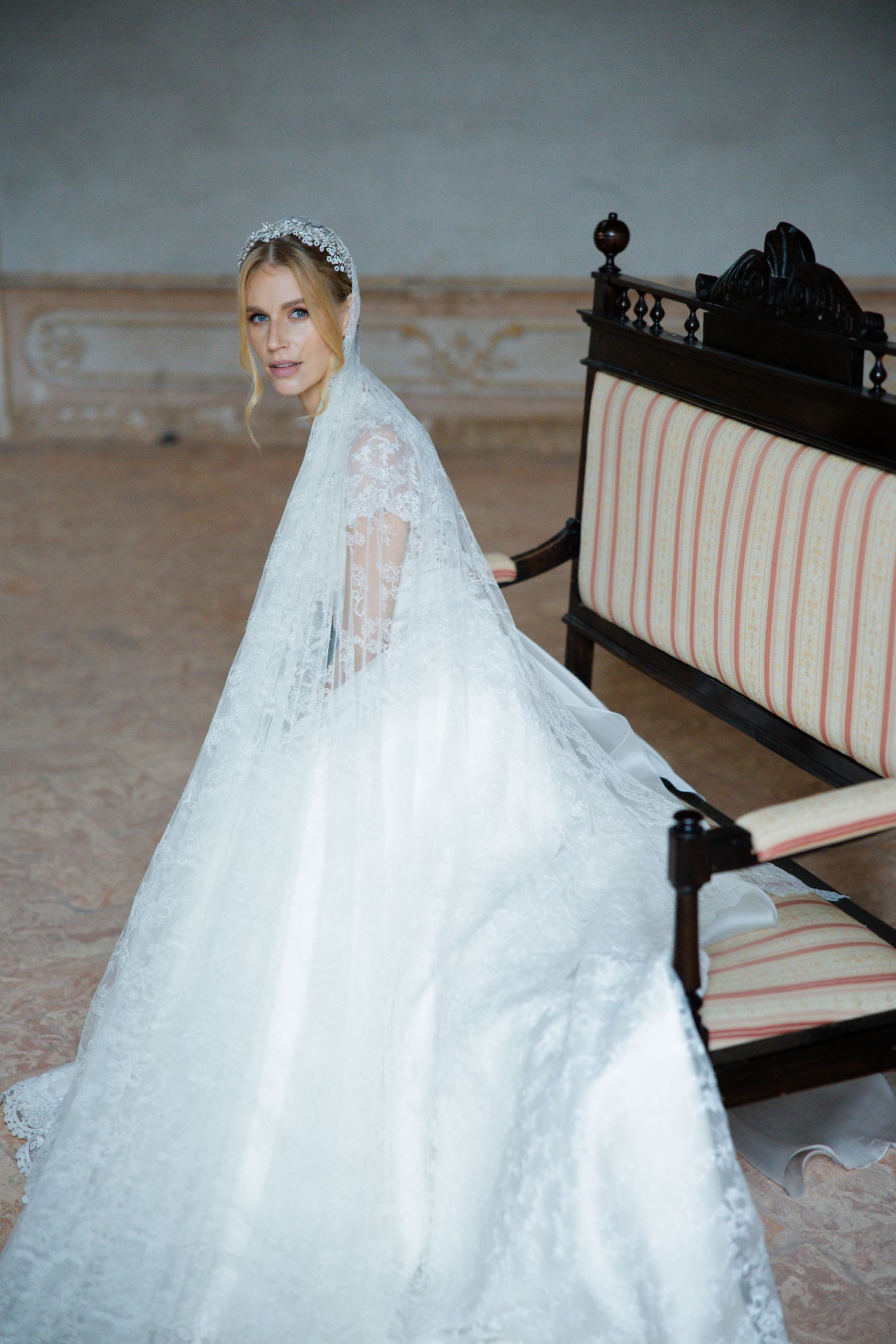Elegant wedding dress with lace veil and ornate train shown against vintage striped bench in moody setting.