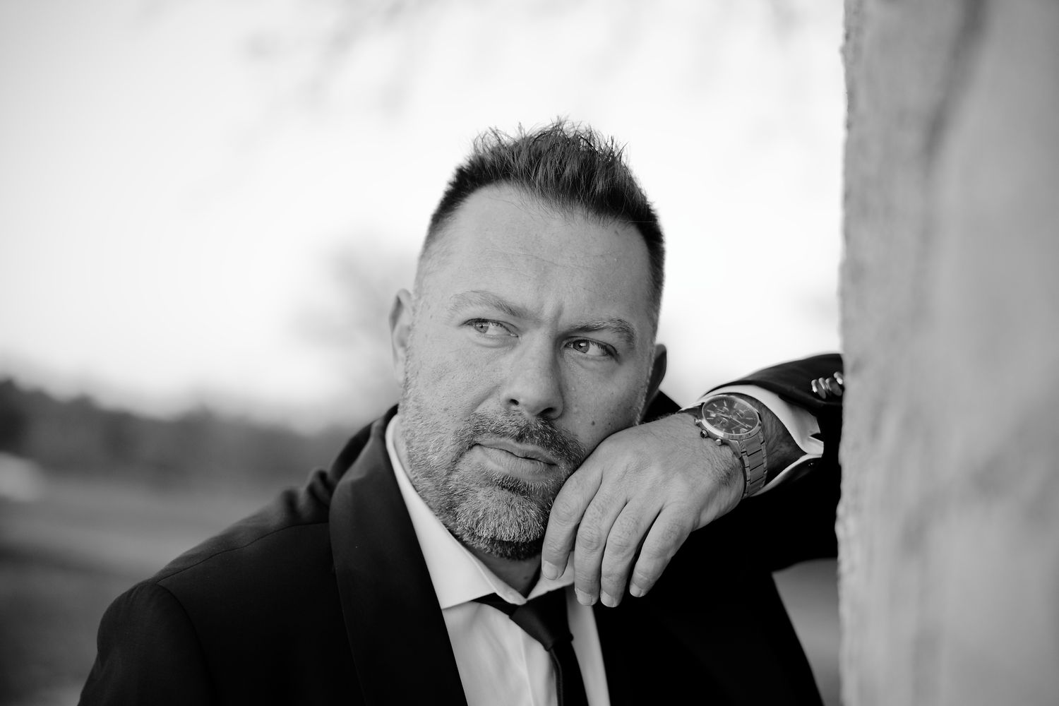 Black and white portrait of someone in a dark suit and tie, resting their hand near their face.