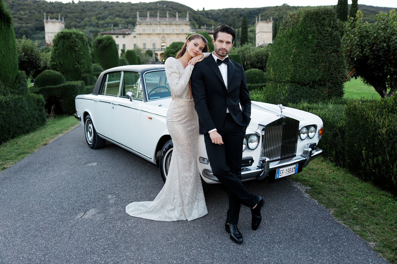 Classic white Rolls Royce parked on driveway with couple in formal attire posing beside luxury mansion and manicured gardens.