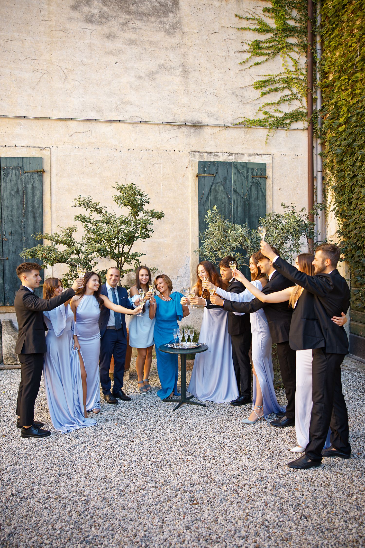 Wedding party in light blue dresses and dark suits celebrating in a courtyard with gravel and stone walls.