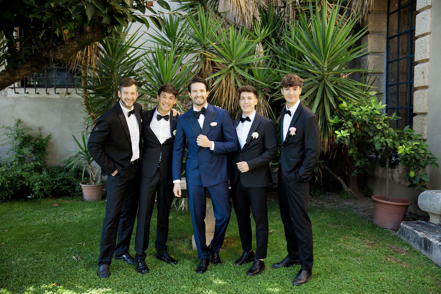 Five groomsmen in tuxedos pose together on a manicured lawn with tropical plants.