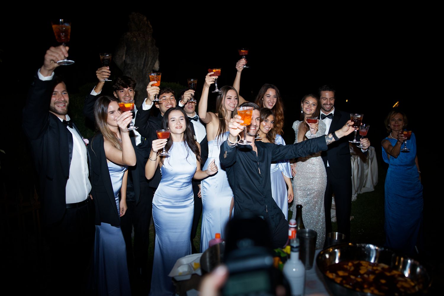 Large group of wedding guests toasting and celebrating at nighttime outdoor reception.