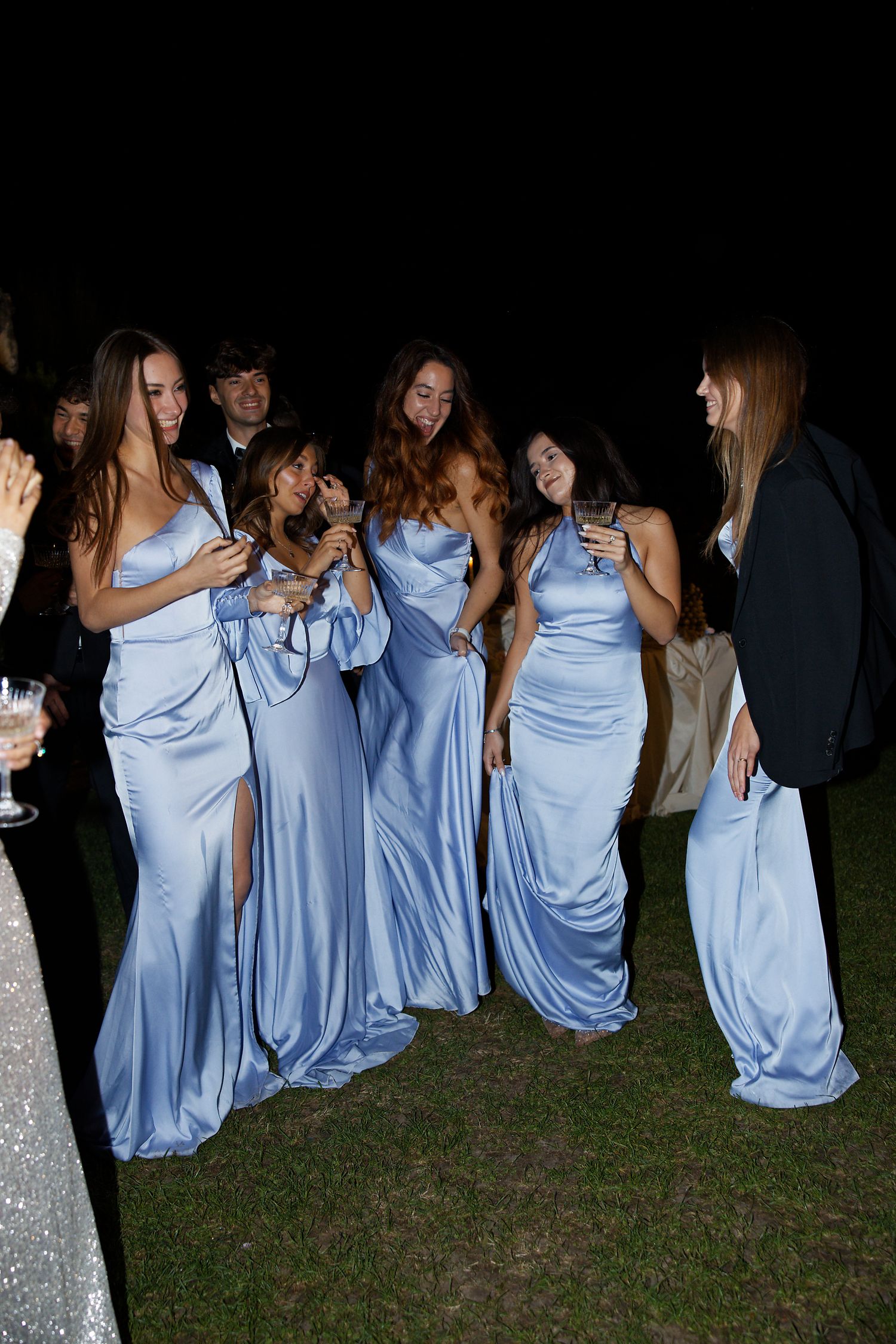 Bridesmaids in flowing light blue dresses share laughs and drinks during an outdoor evening celebration.
