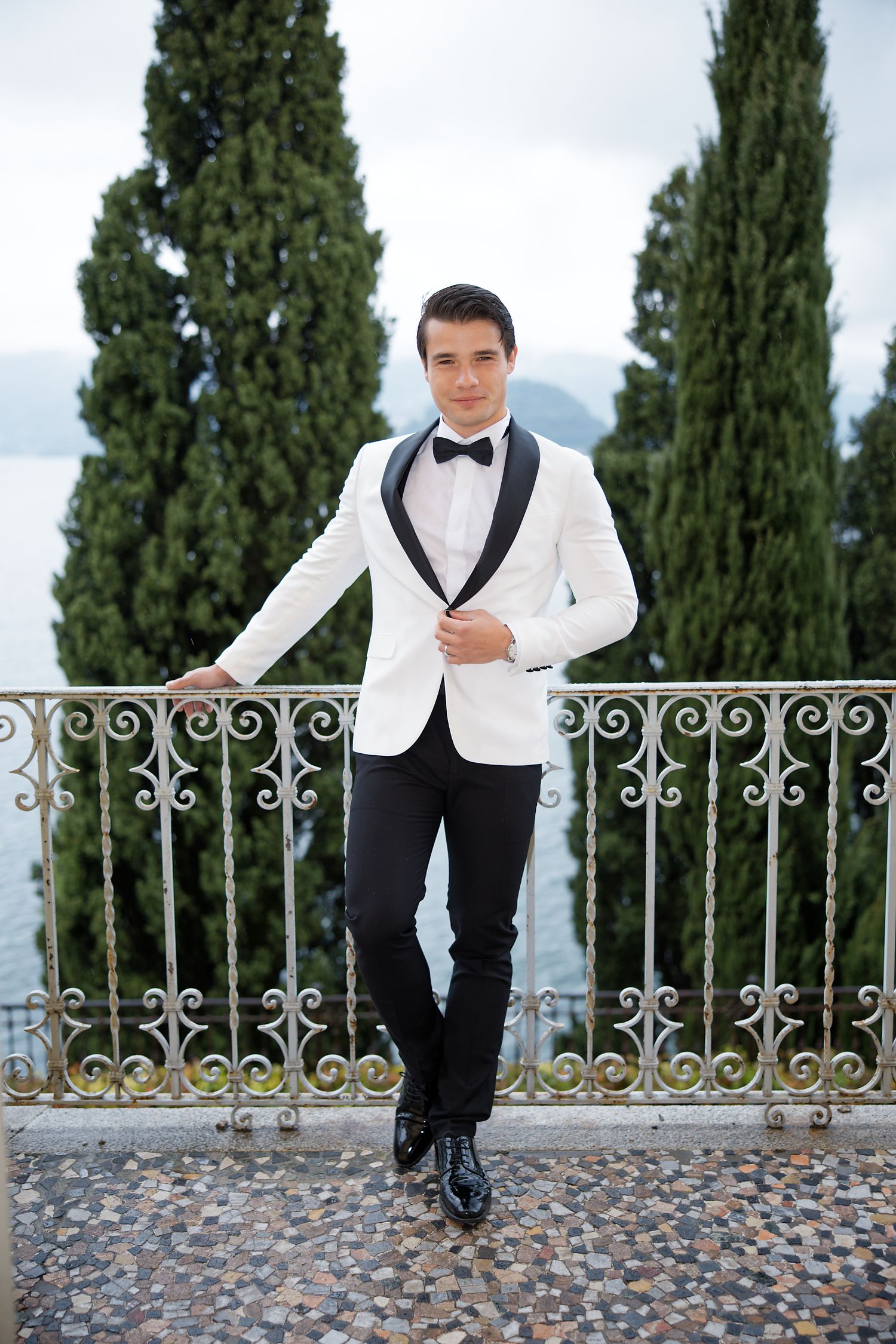 A groom in a white dinner jacket poses on a stone balcony overlooking a lake.