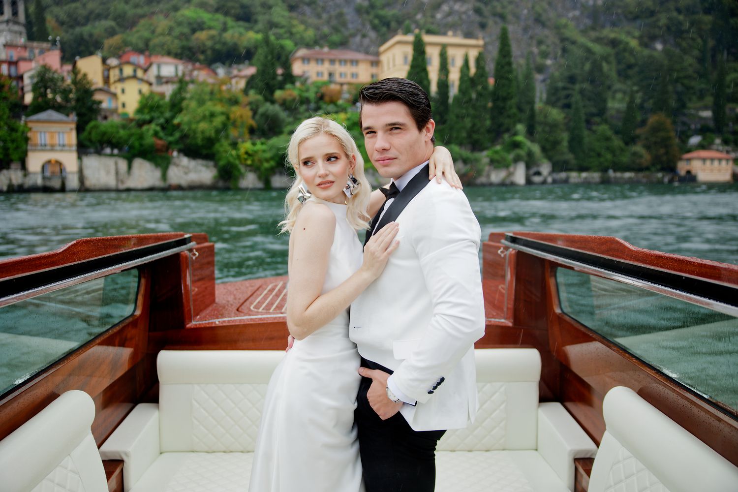 A newlywed couple poses on a wooden boat with white seating against a scenic Italian lake backdrop.