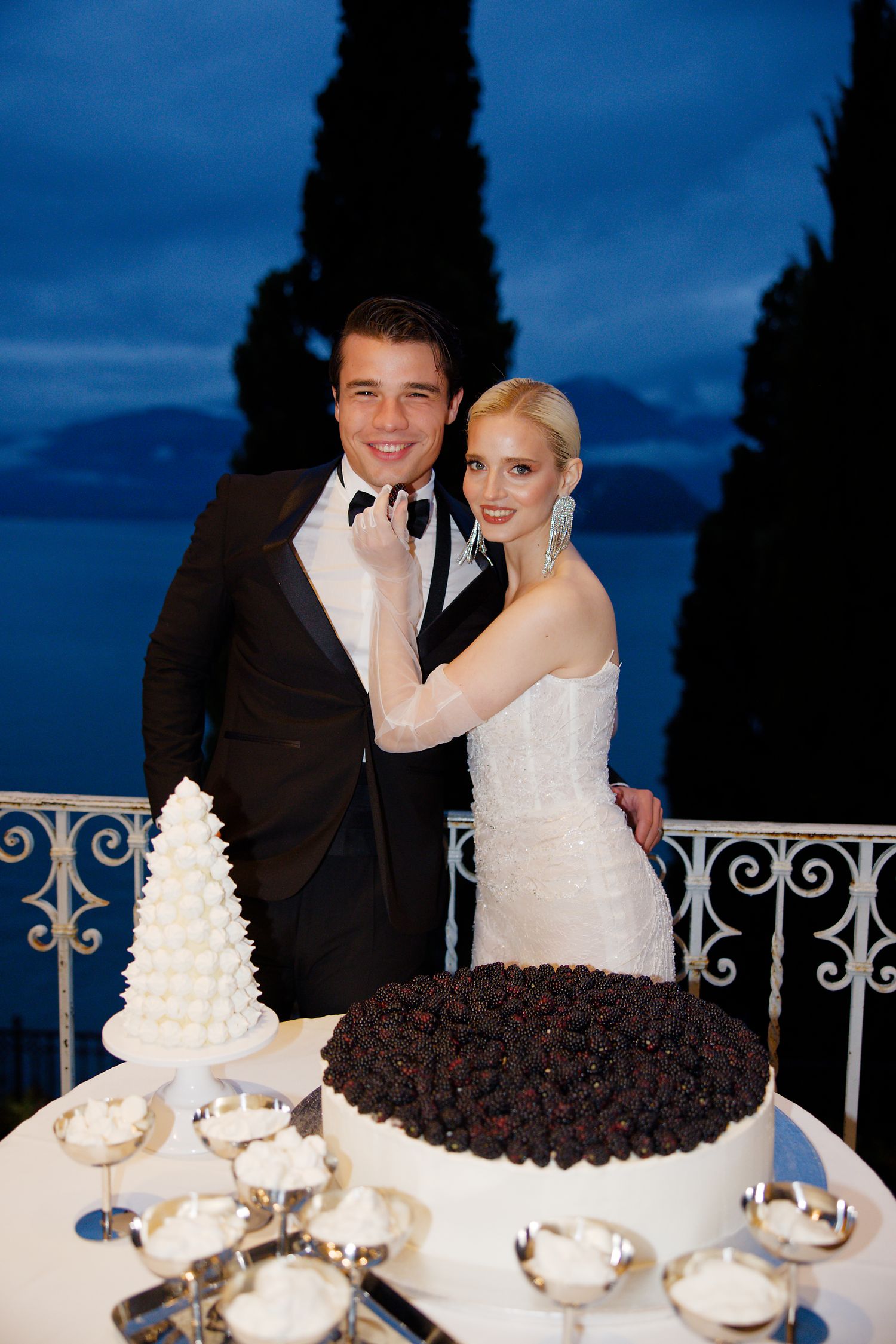 Newlyweds pose by their wedding cakes on balcony at dusk with cypress trees and lake in background.