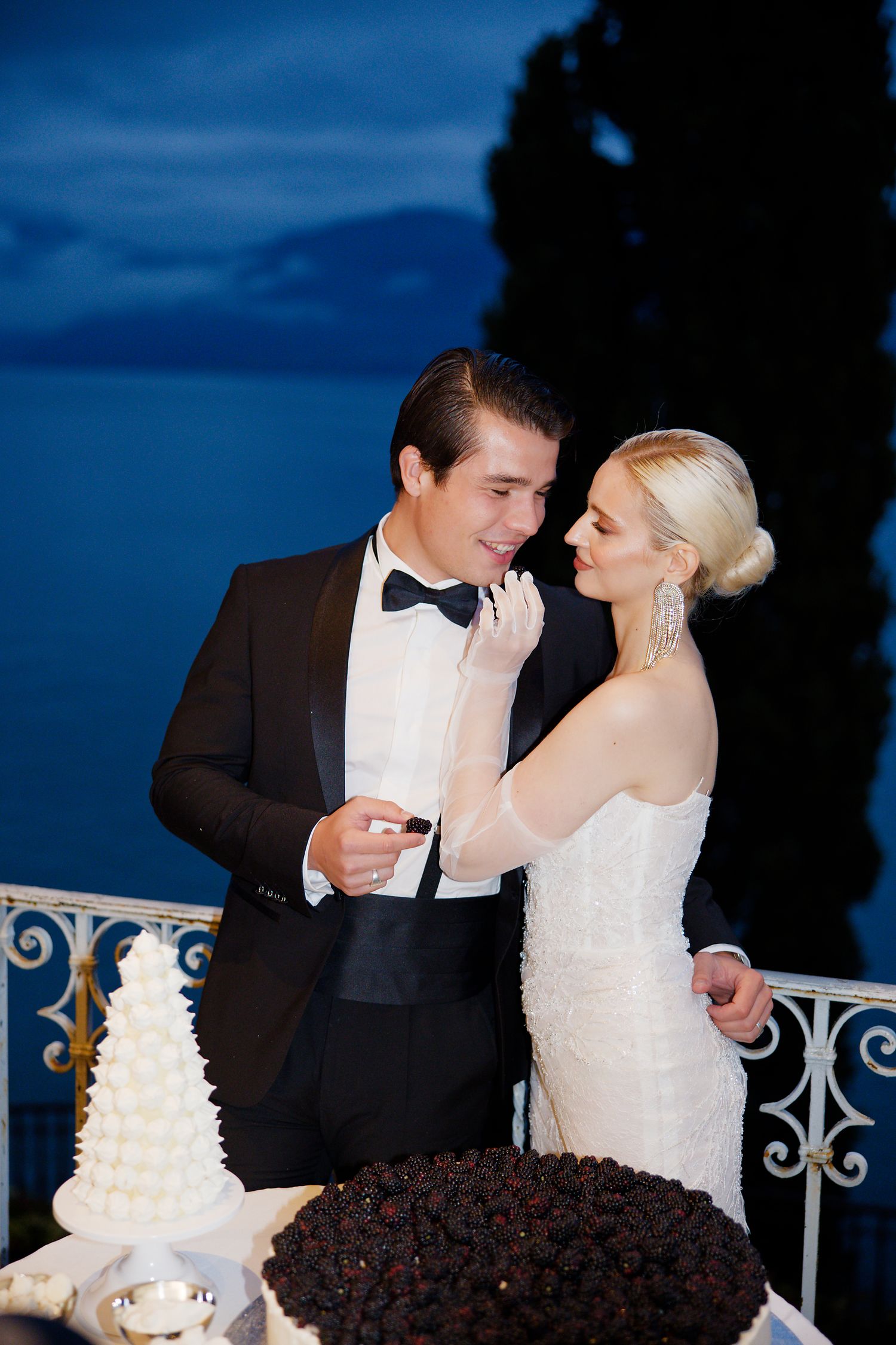 Elegant wedding couple shares an intimate moment by their wedding cakes at twilight on a balcony.