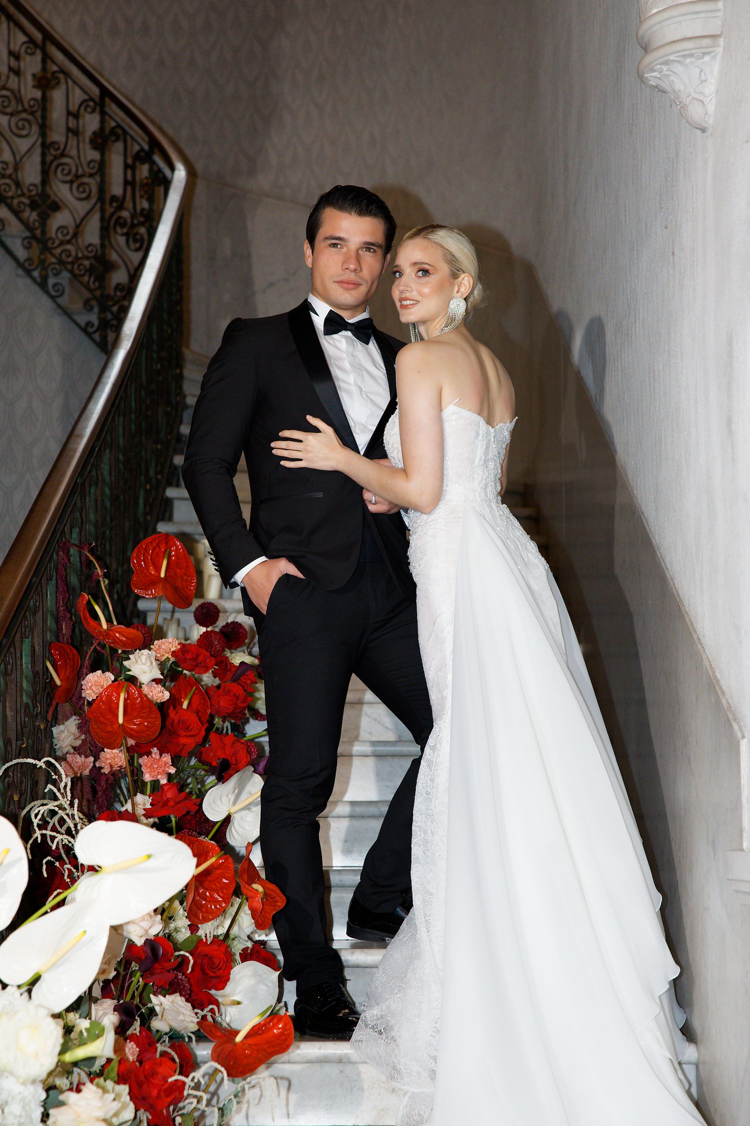 A newlywed couple poses on elegant white stairs decorated with vibrant red and white floral arrangements.