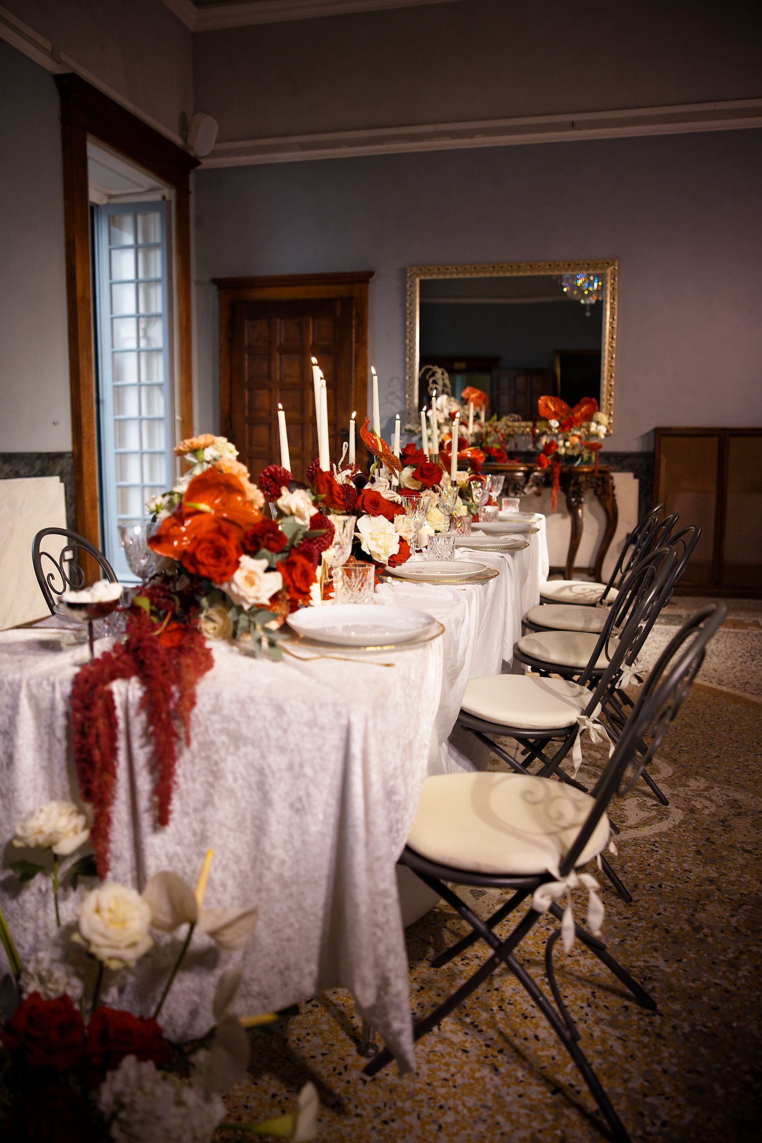 Elegant wedding reception table with white linens, taper candles, and vibrant red and orange floral arrangements.