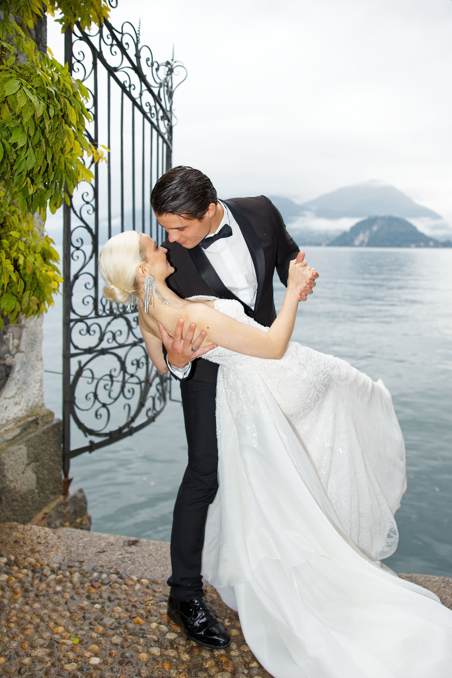 Dramatic dip pose against ornate gates with lake and mountains in background.