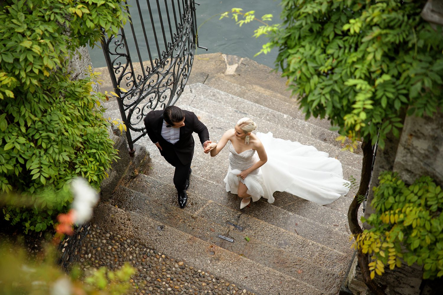Couple descending ornate stone steps hand in hand at lakeside garden.