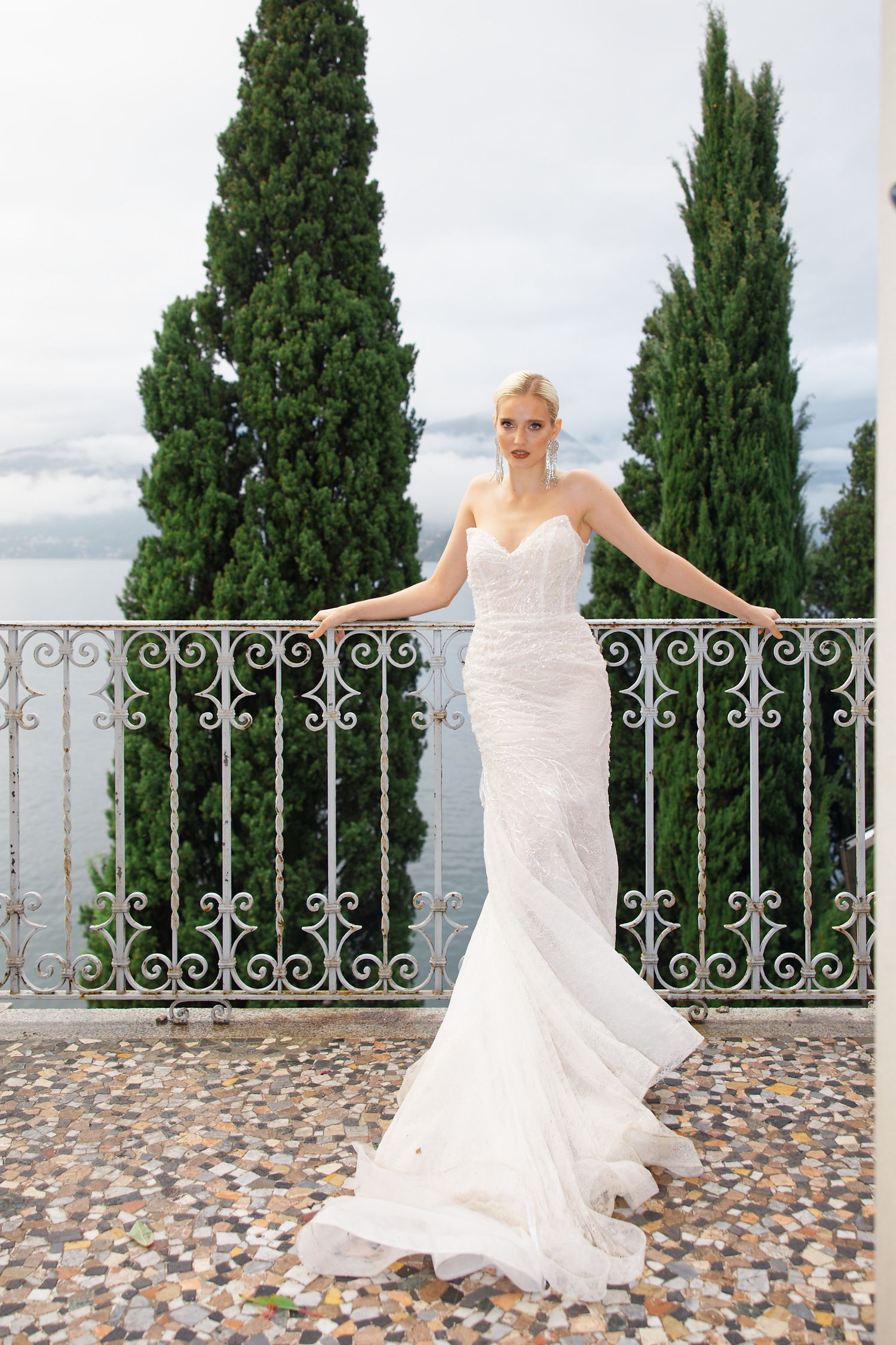 Stunning bride in fitted lace gown poses on stone terrace with cypress trees and lake view.