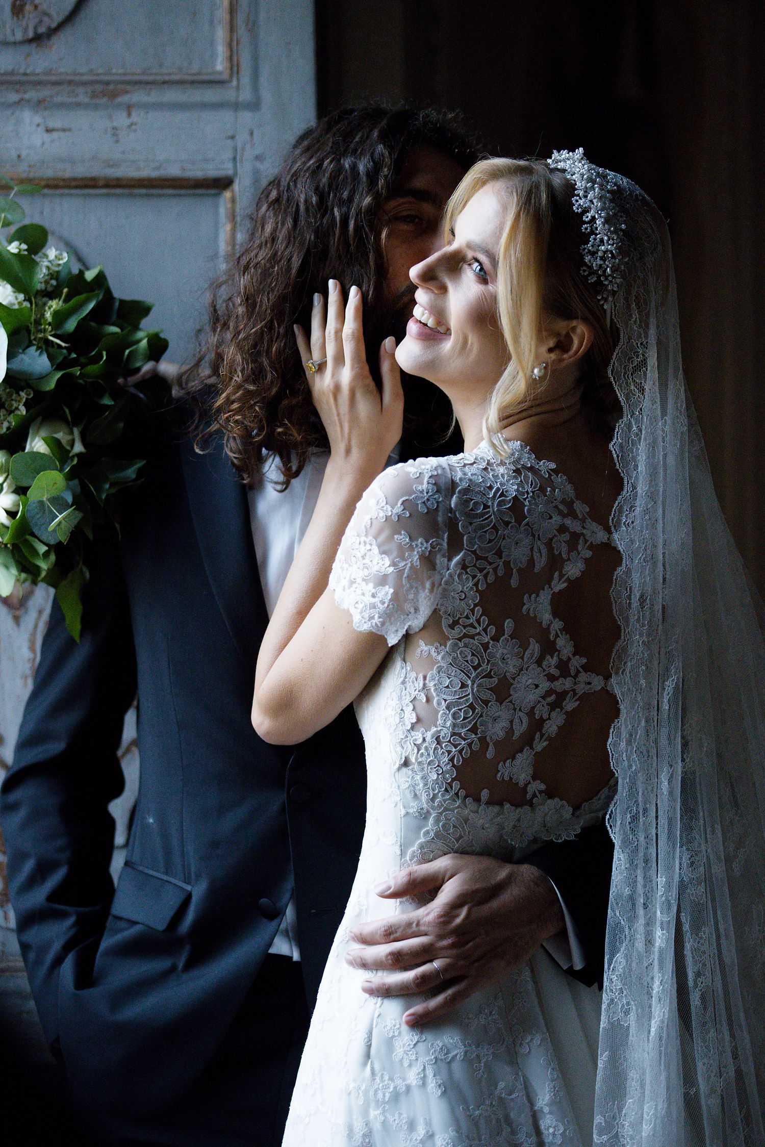 An intimate wedding moment captured against a vintage doorway with intricate lace details and greenery bouquet.