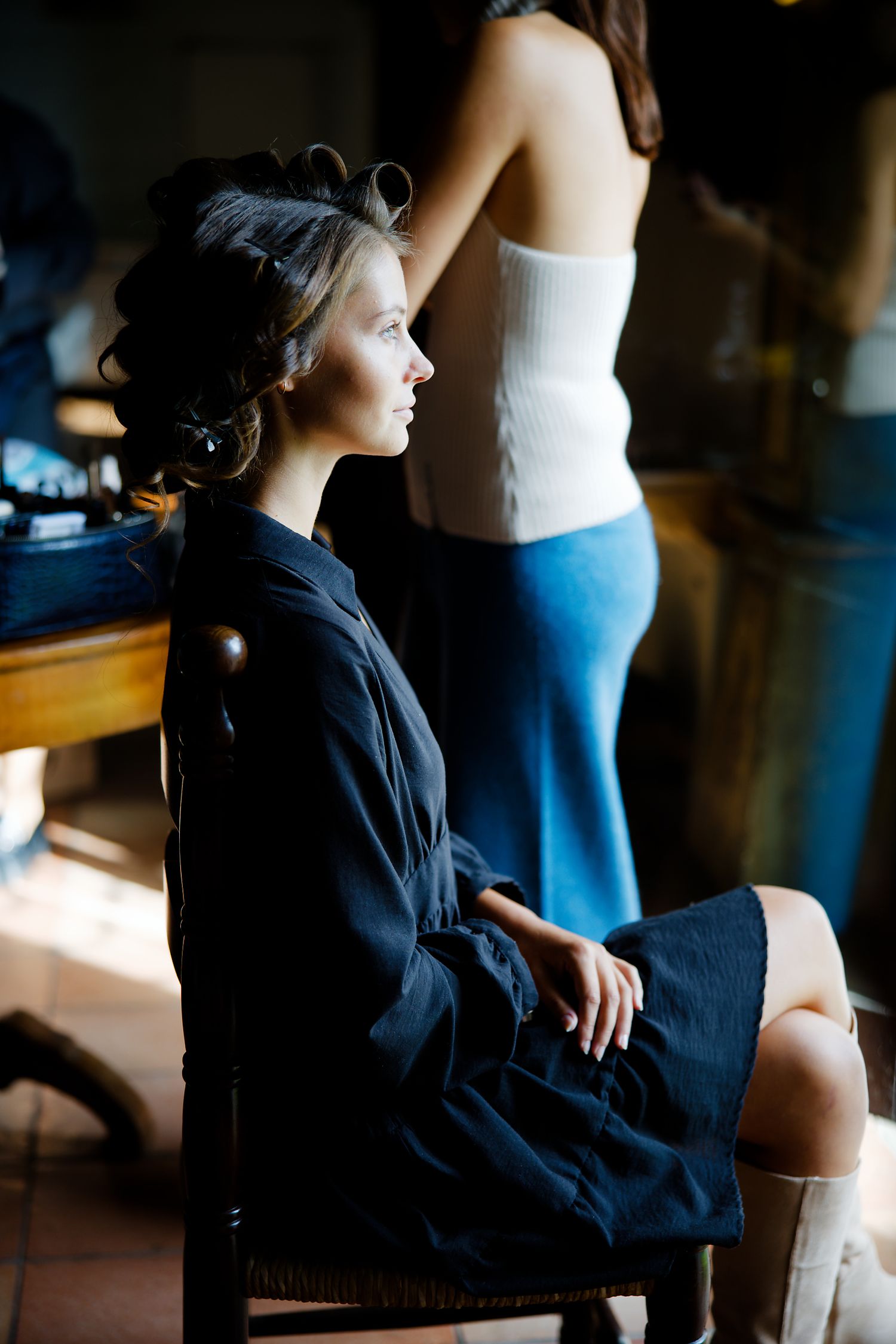 Person getting hair and makeup done in a black silk robe while seated in a vintage wooden chair.