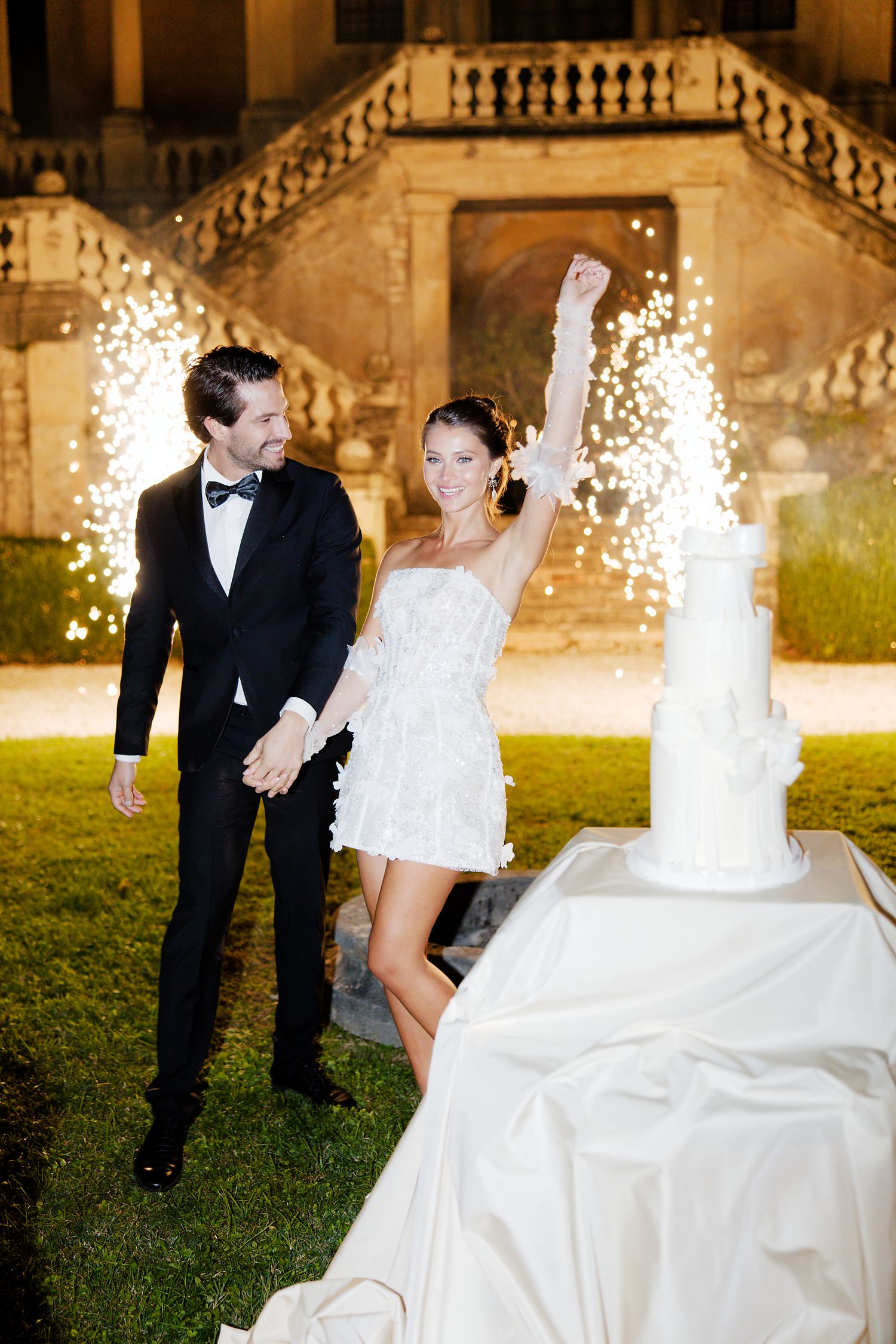A celebratory moment with sparklers and a wedding dress train on the lawn of a majestic villa at night.