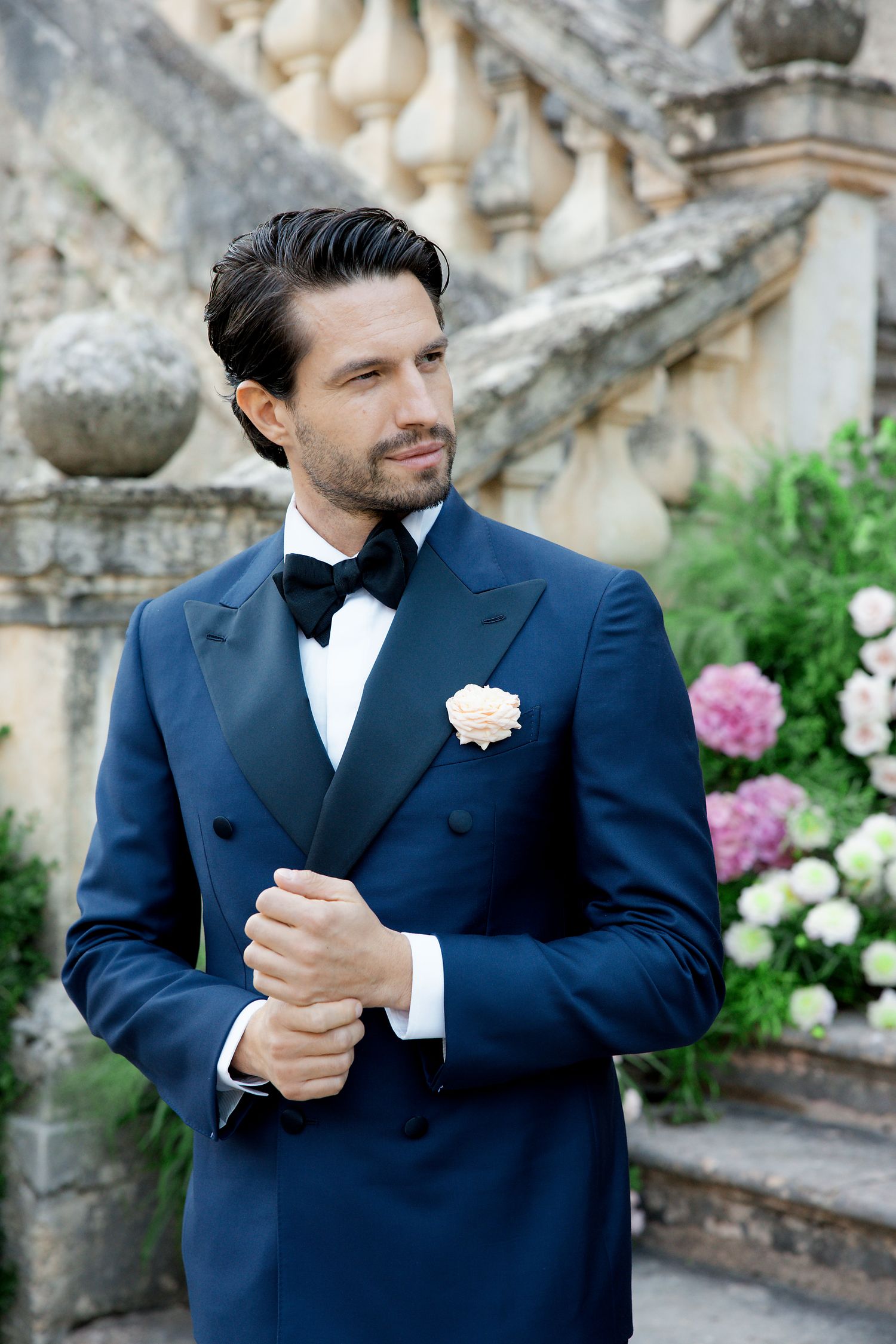 A groom in a navy blue tuxedo with white boutonniere poses against an ornate stone staircase with blooming flowers.