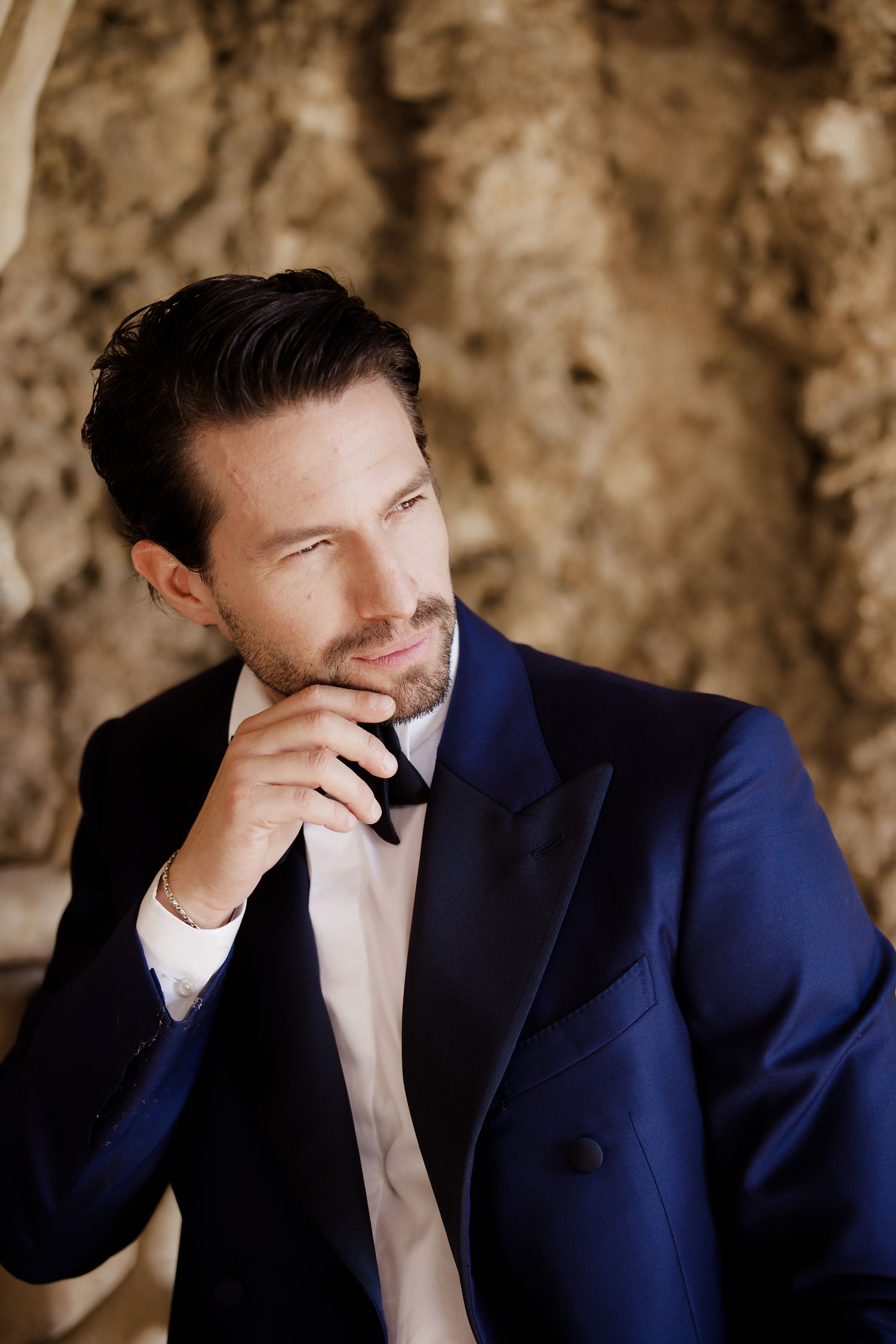 A man in a navy tuxedo and bowtie poses thoughtfully against a textured stone wall.