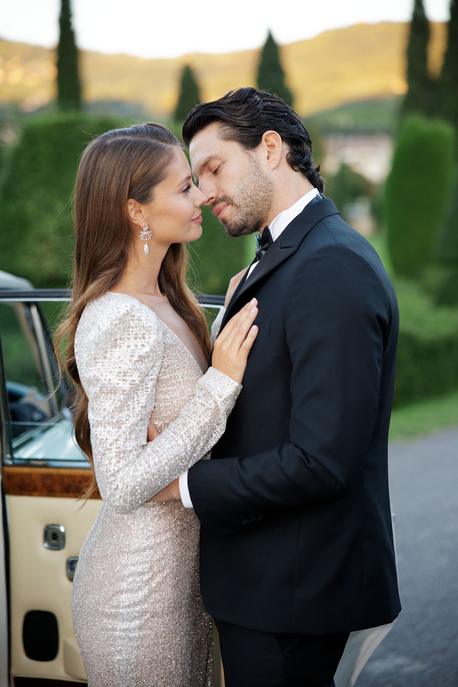 A tender embrace between a couple in formal wear standing beside a classic car.