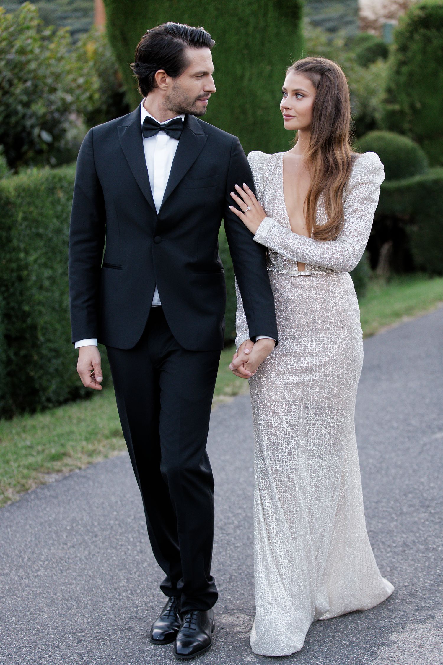 Elegant couple in formal wear walking together on garden path at dusk.