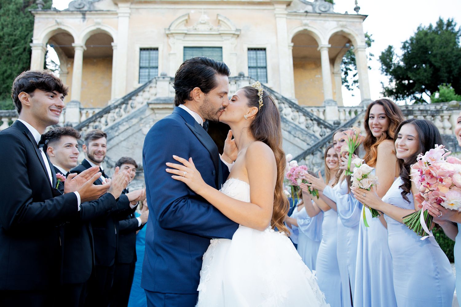A crowd of wedding guests celebrates as newlyweds share a kiss on grand villa steps.