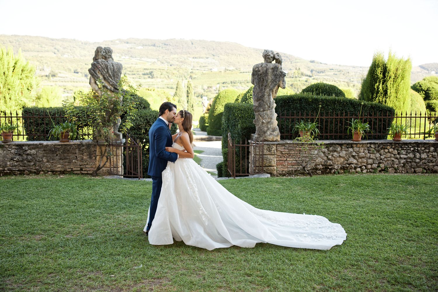 Romantic wedding couple embrace in an elegant garden setting with stone sculptures and rolling hills in the background.