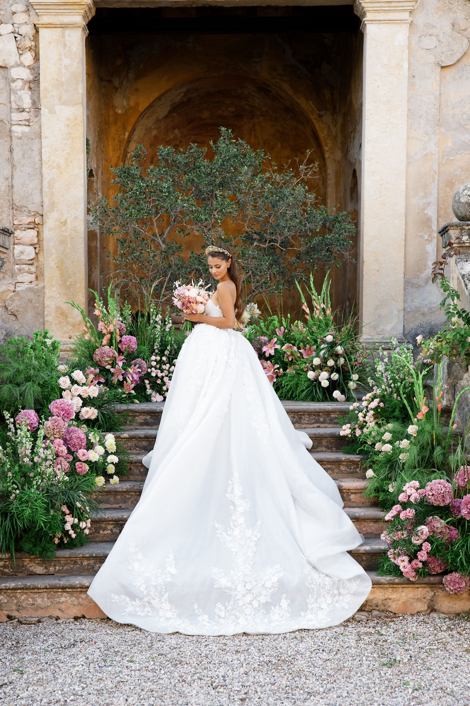 A wedding dress with long train photographed against stone architecture and floral arrangements.