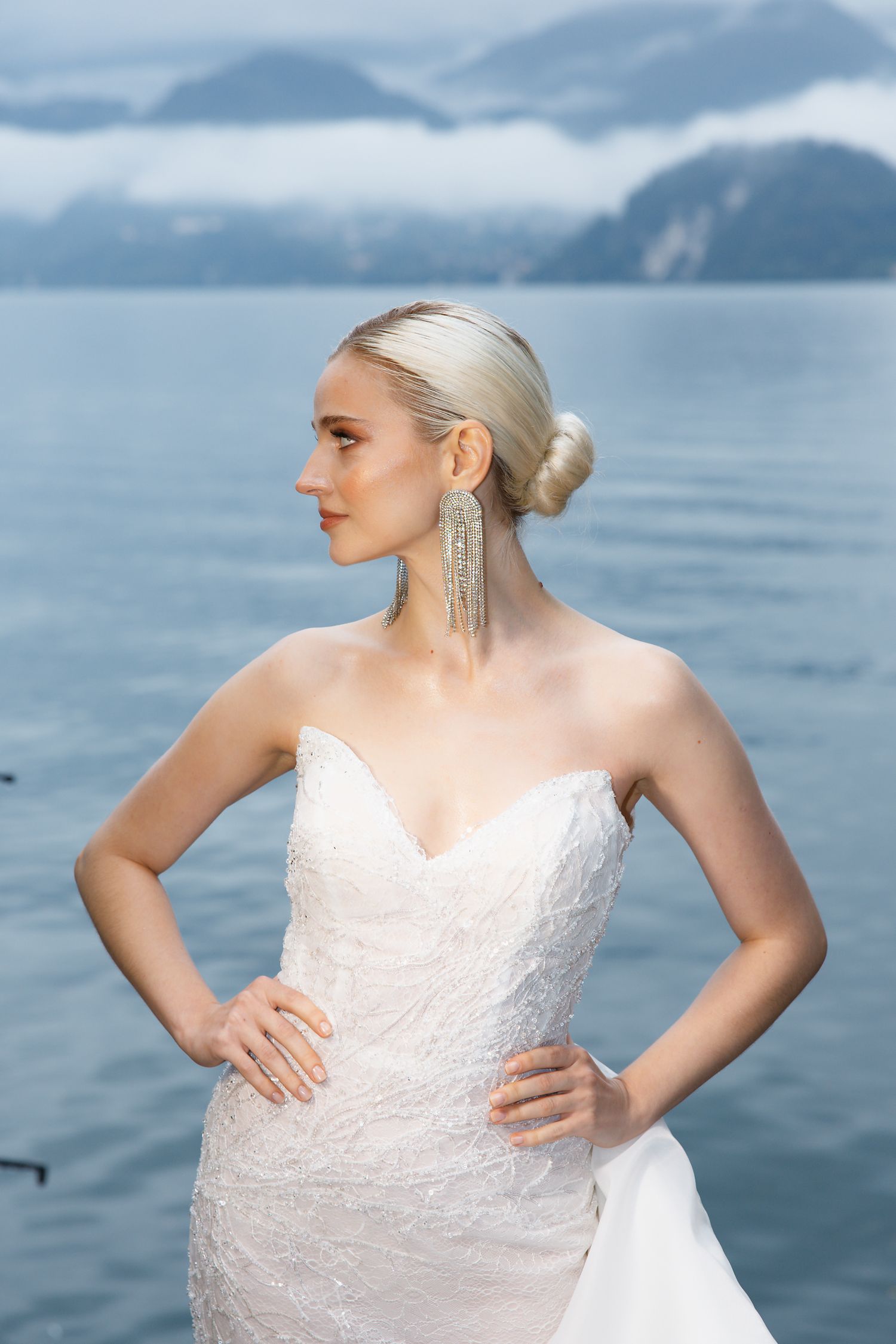 Elegant bride in white gown poses against mountain lake backdrop.