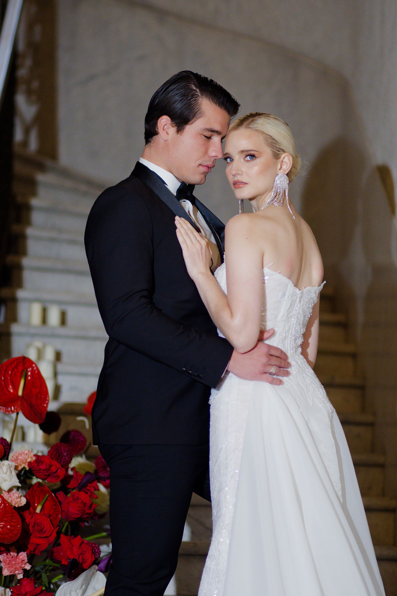 Romantic couple embraces on grand staircase with red roses, bride in white gown and groom in tuxedo.