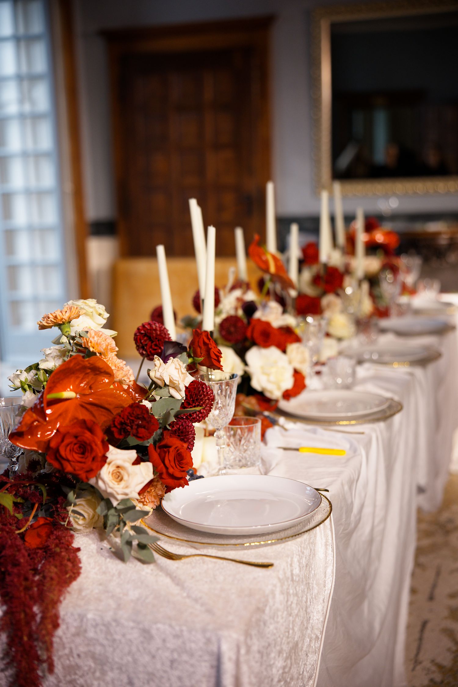Wedding table setting with white plates, tall candles, and dramatic red and orange flower centerpieces.