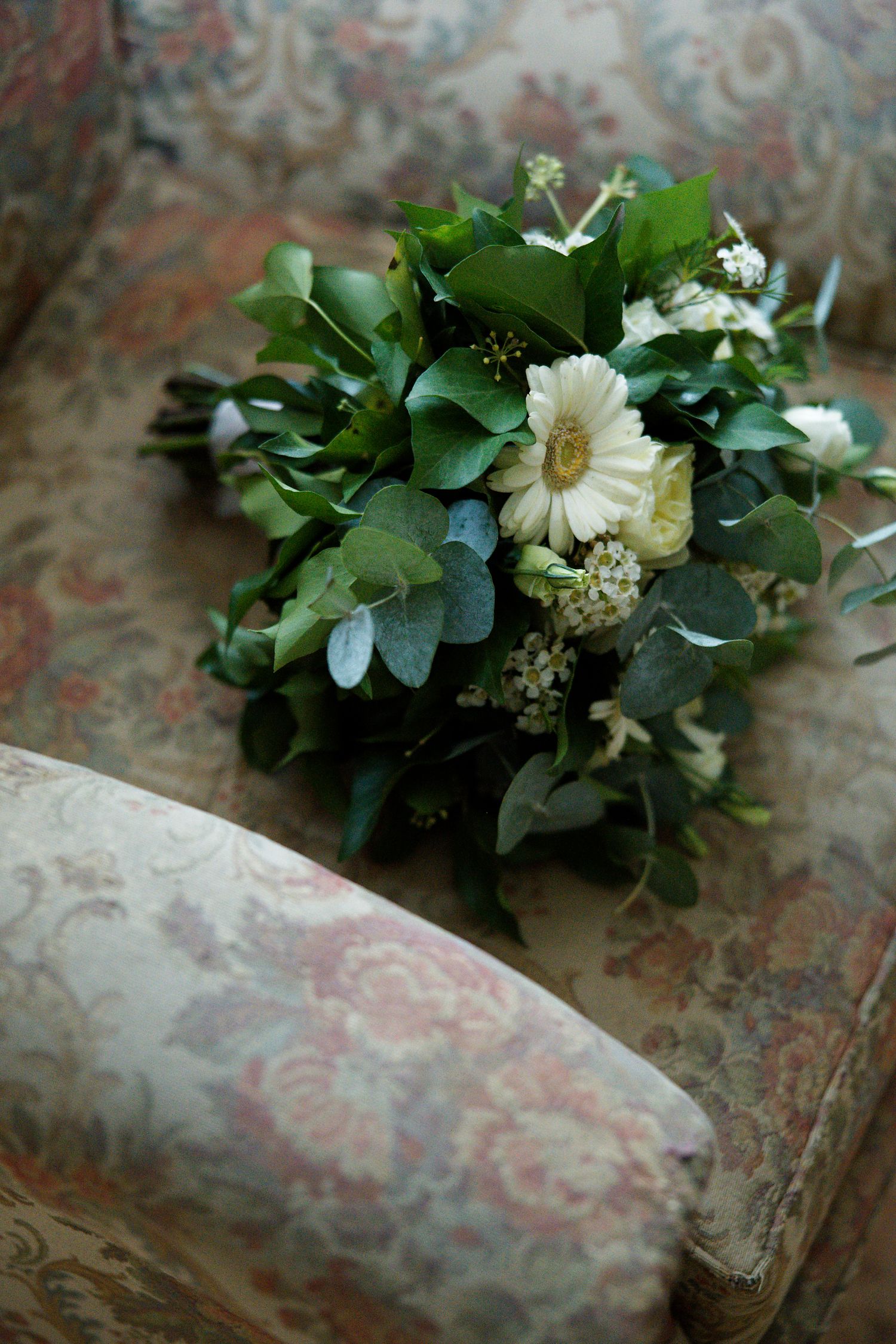 A delicate wedding bouquet with white daisies and lush green foliage rests on a floral patterned surface.