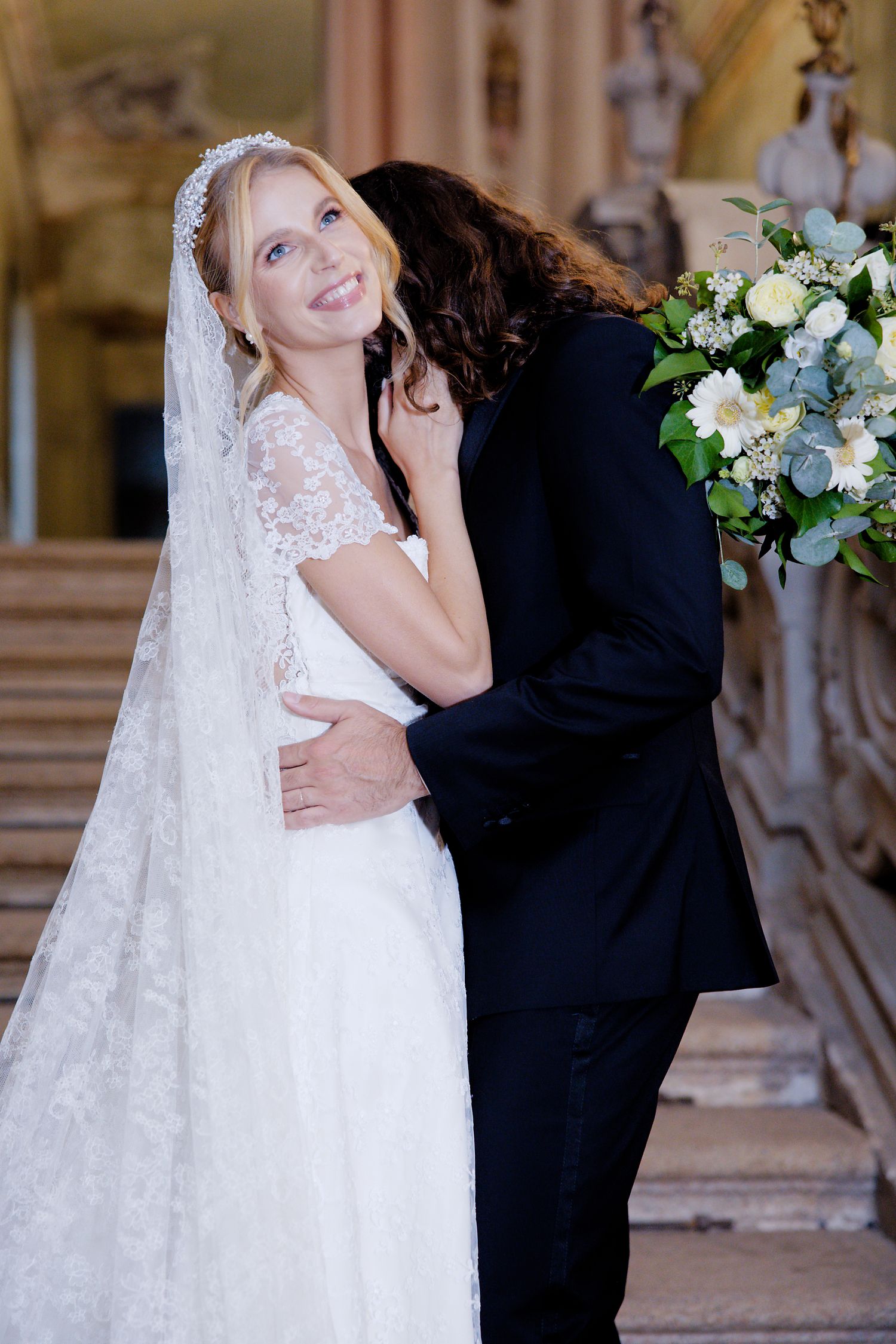 A romantic wedding moment captured in an ornate venue with a beautiful lace dress and elegant bouquet of white flowers.