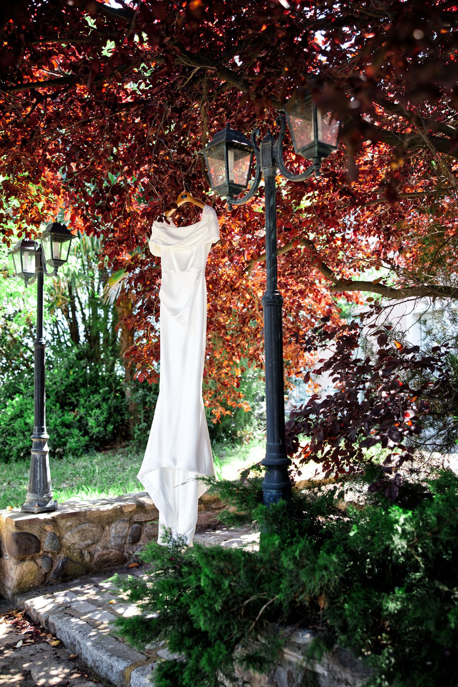 An elegant white wedding dress hangs from a vintage lamppost against vibrant red autumn foliage in a garden setting.