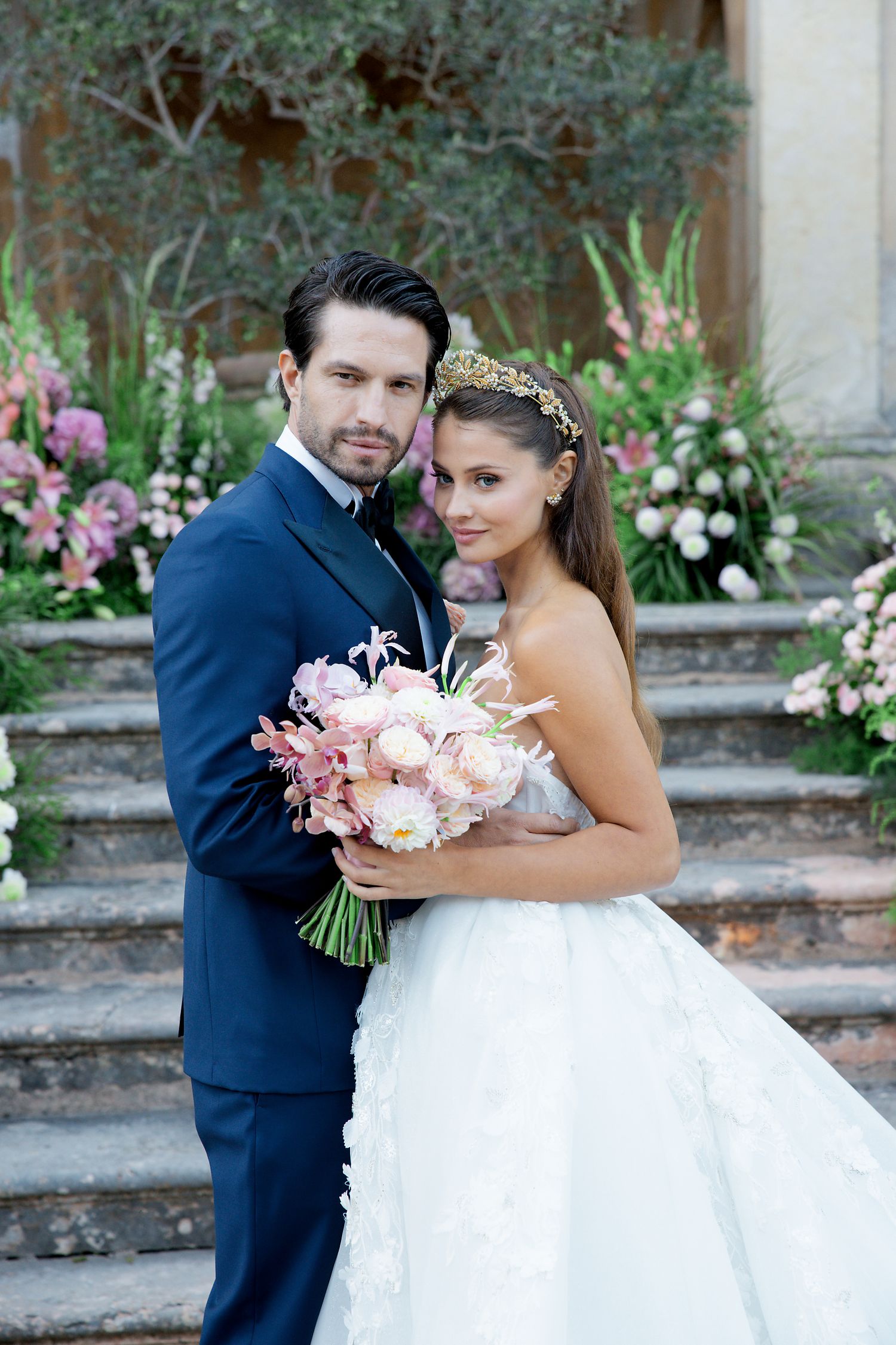 Elegant wedding couple poses on stone steps with pink floral bouquet and garden backdrop.