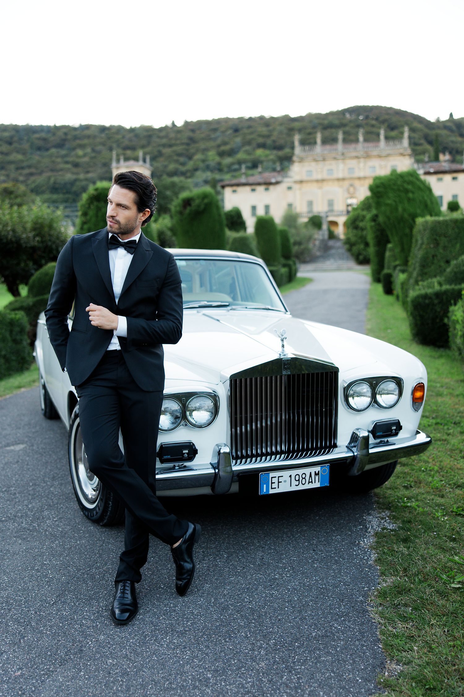 A well-dressed person poses against a classic white Rolls Royce with an Italian villa in the background.