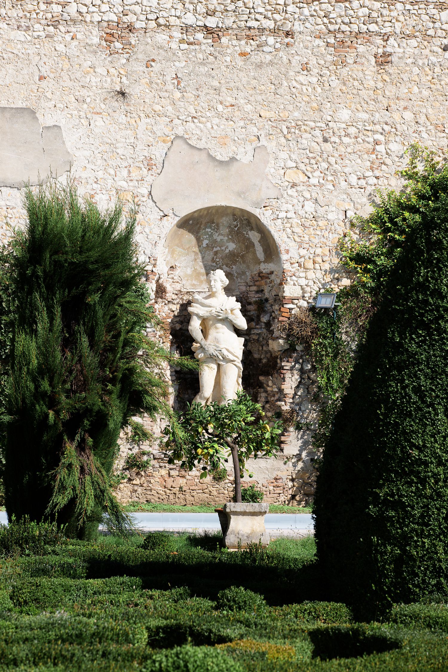Classical marble statue in garden alcove surrounded by manicured shrubs and stone walls.