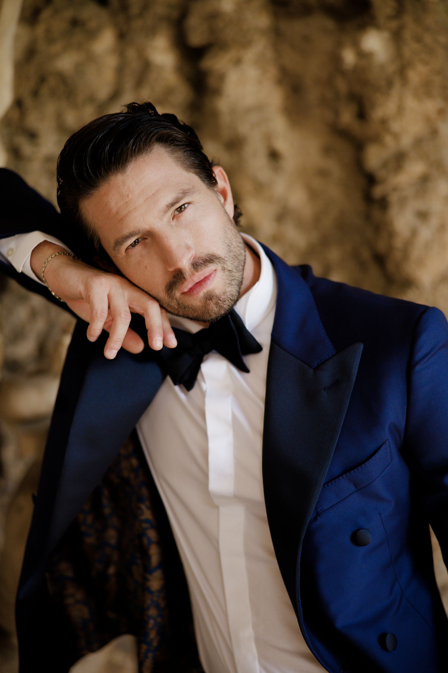 A dapper gentleman in a navy tuxedo leans against a stone wall in a pensive pose.