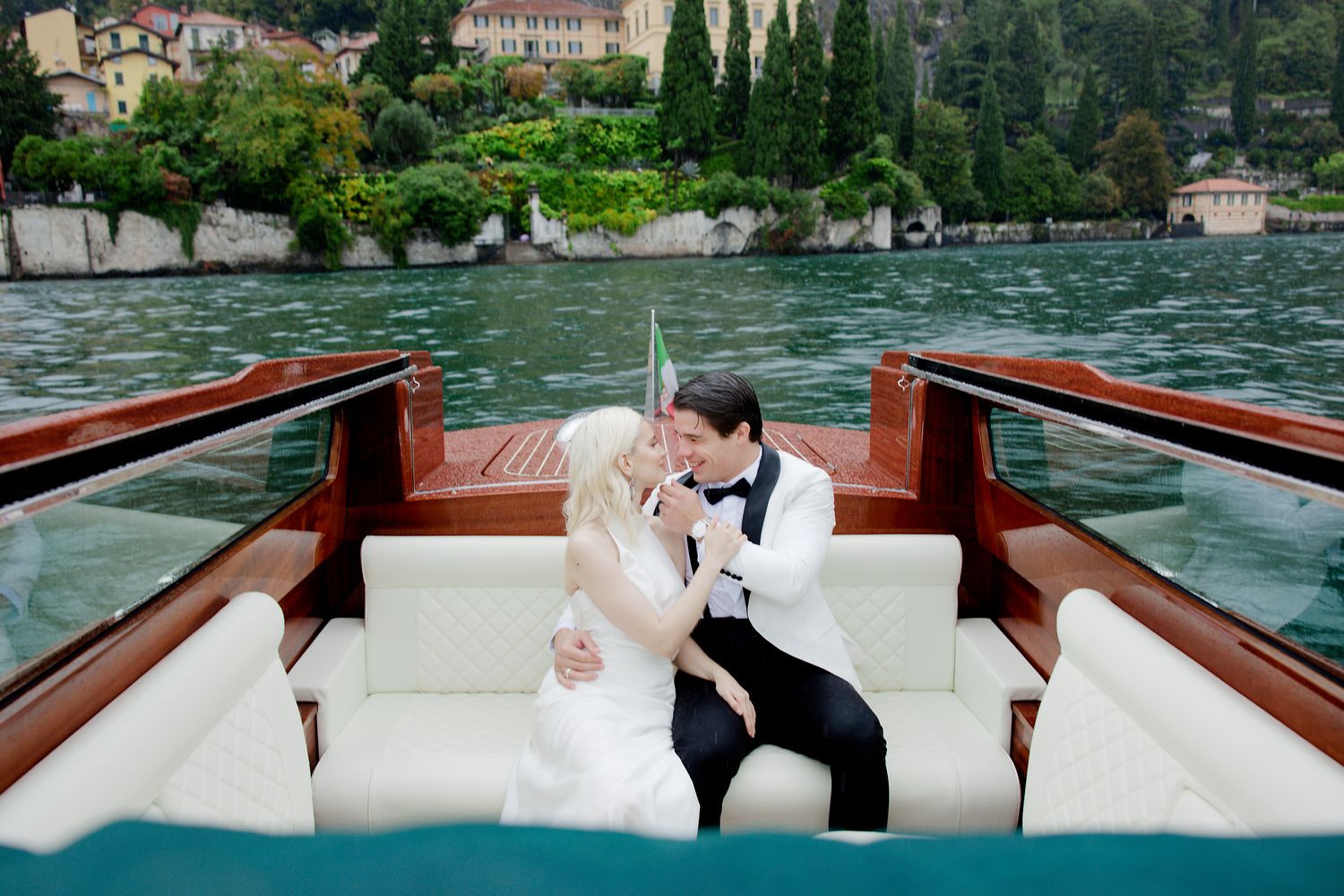Wedding couple embracing on classic wooden boat against Mediterranean coastal backdrop.