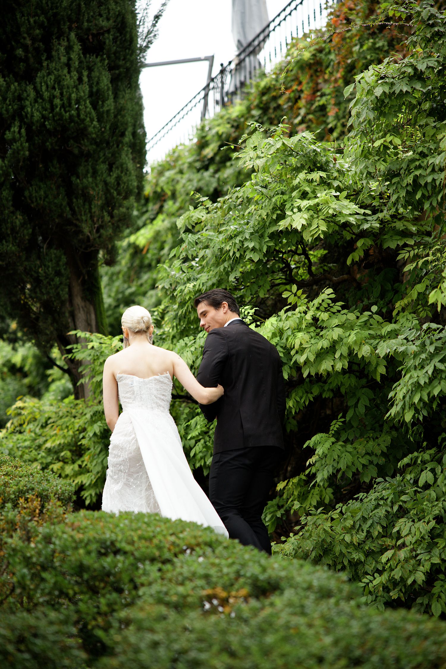 A couple in wedding attire walks through a lush garden path.