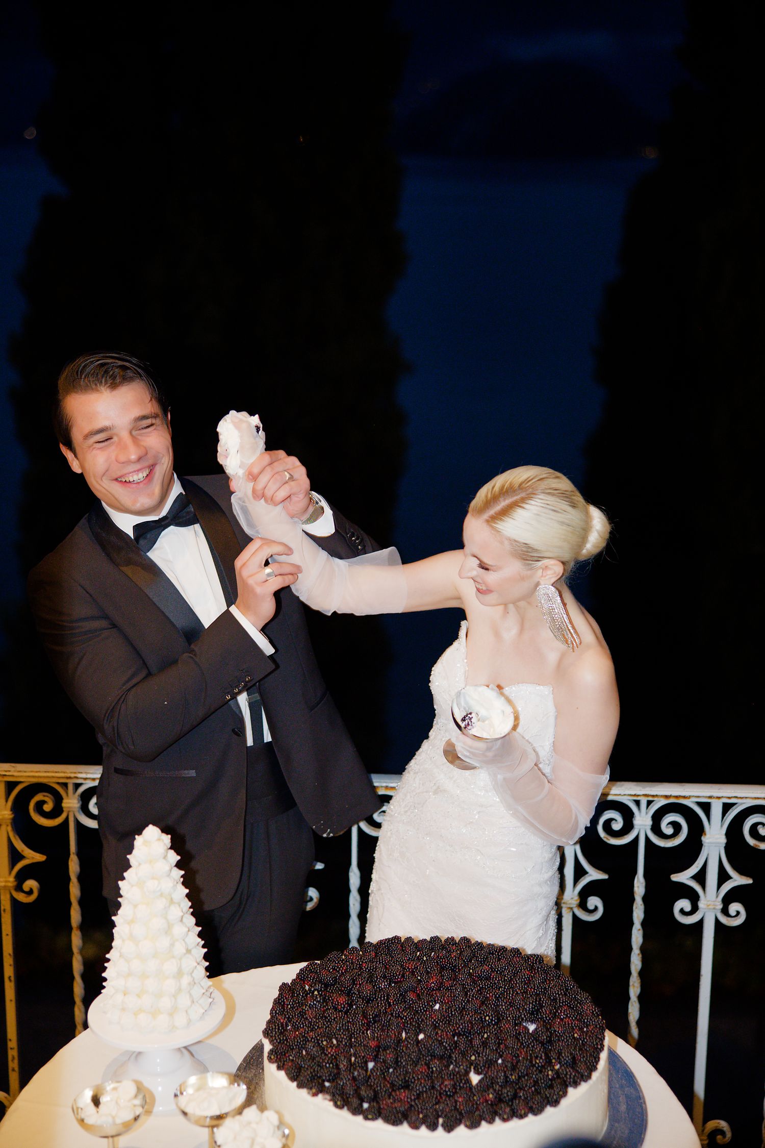 Wedding couple playfully shares cake at elegant outdoor reception with ornate railing and lake views.