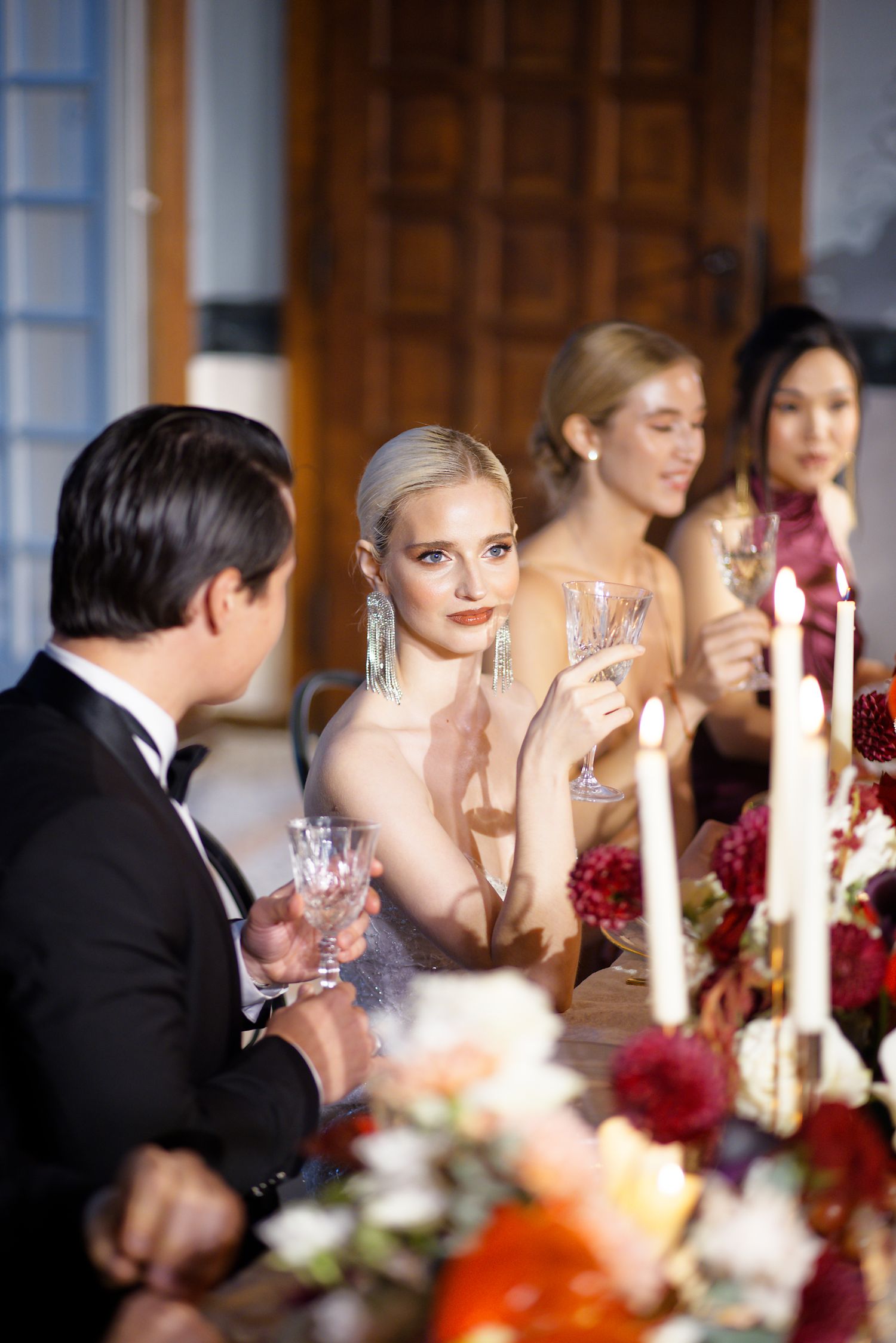 Elegant guests in formal attire enjoying drinks at a luxurious candlelit dinner table with floral centerpieces.