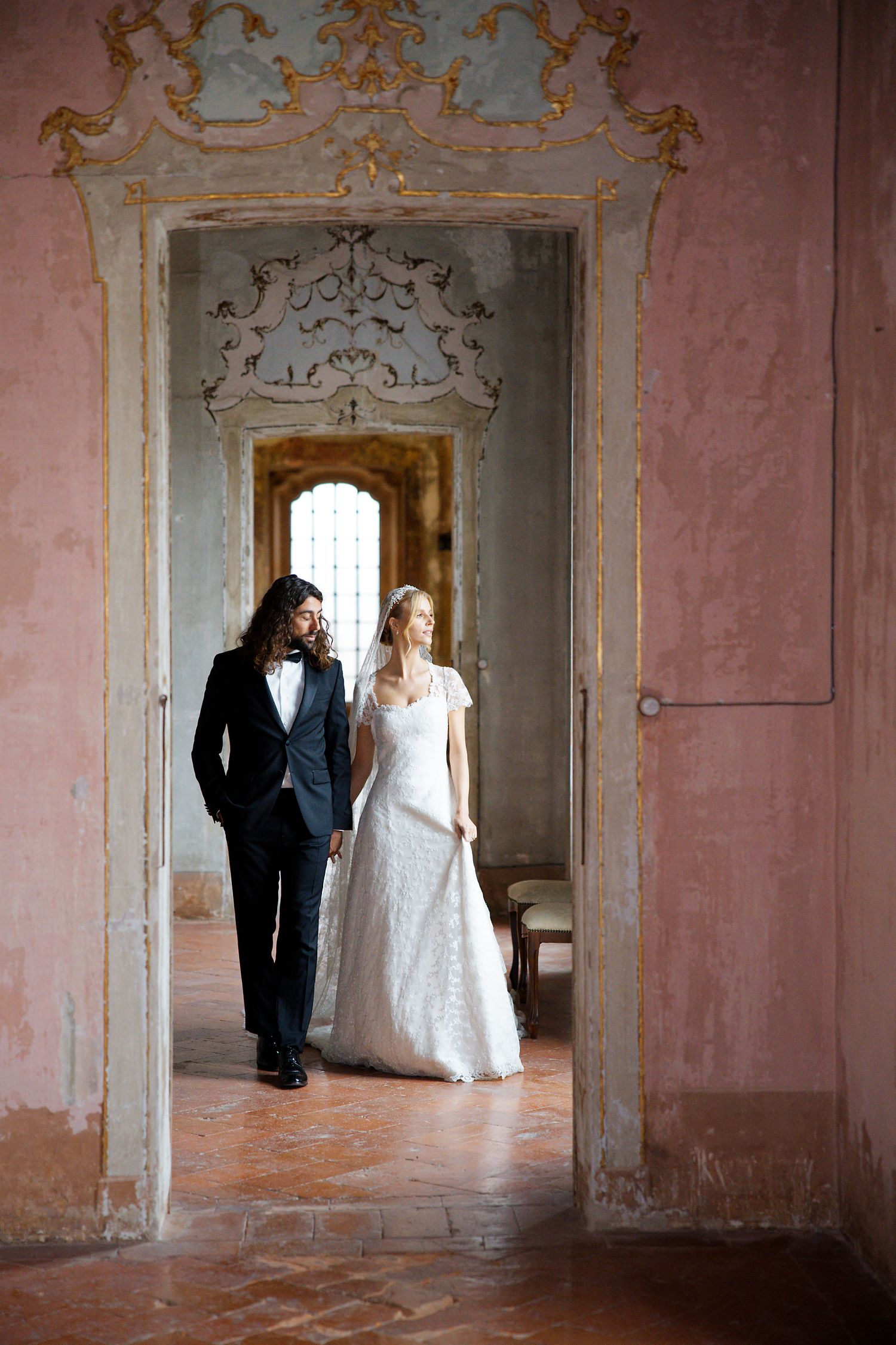 A romantic wedding portrait framed within an ornate doorway of a historic palace with pink walls and baroque details.