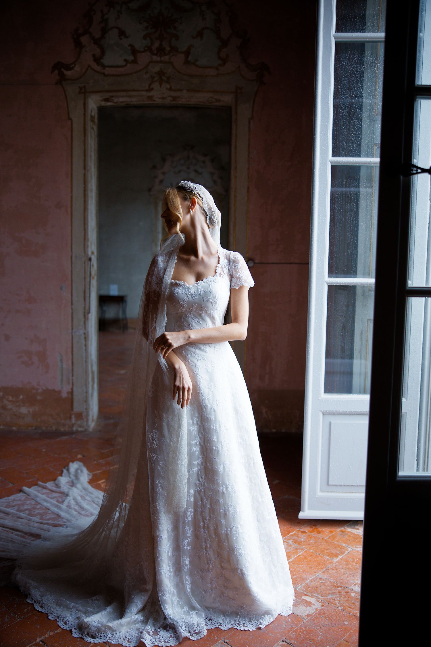 Bride in vintage lace gown with cap sleeves stands by window in historic interior.