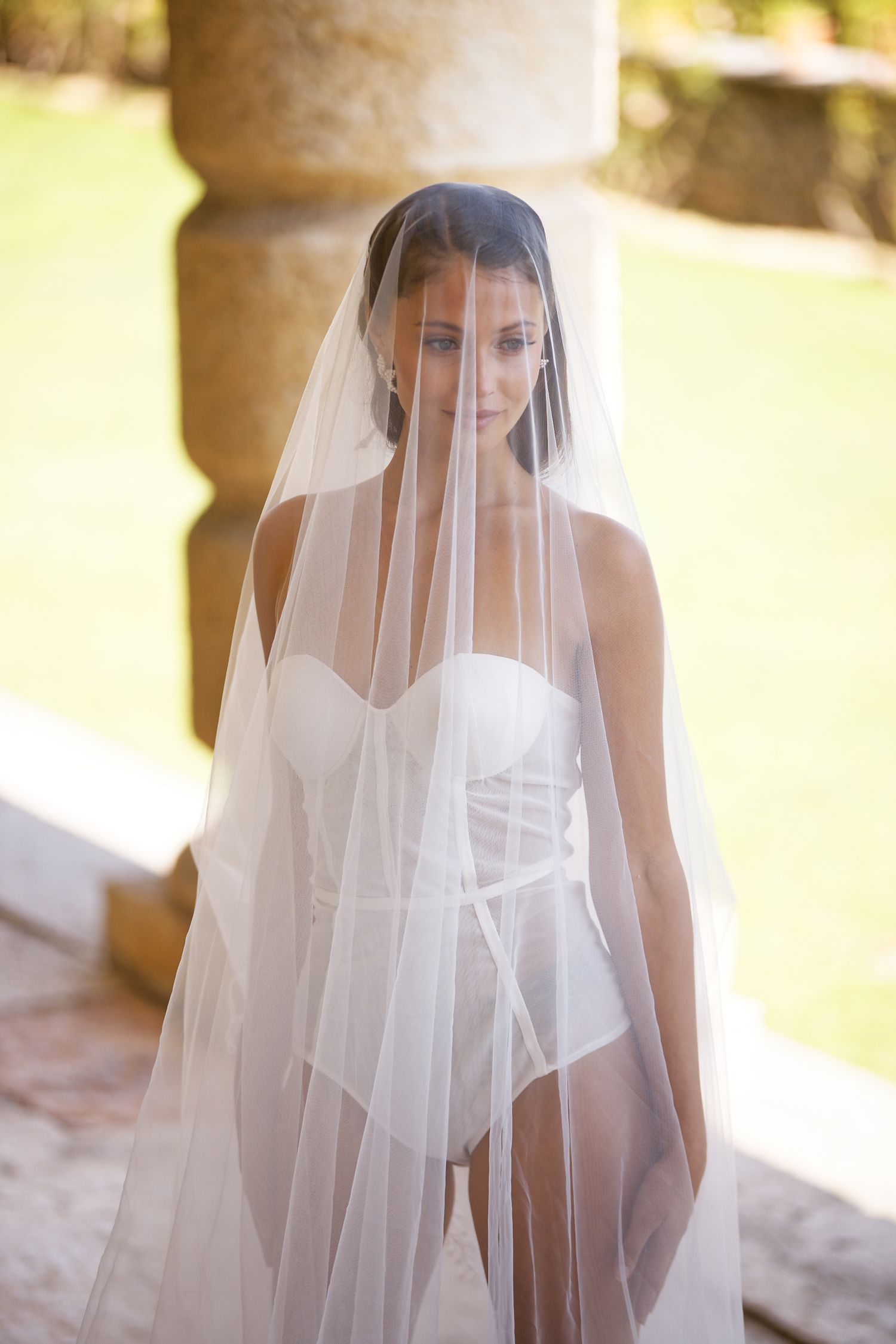 Ethereal bridal portrait with sheer veil and white bodysuit in golden sunlit outdoor setting.