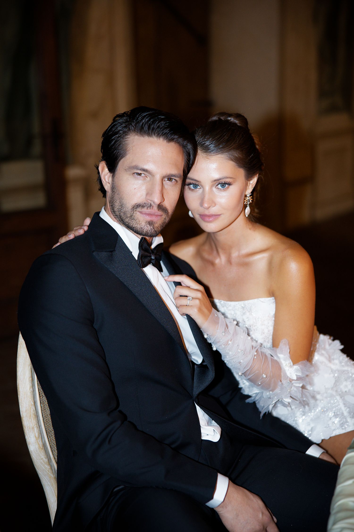 Elegant wedding portrait featuring couple in black tuxedo and white lace gown against classic interior.