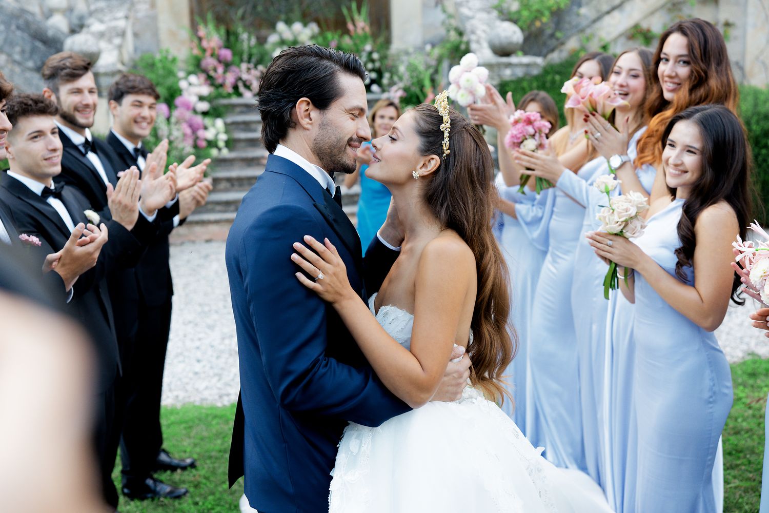 A newly married couple embraces while wedding guests in light blue dresses applaud in an outdoor garden.