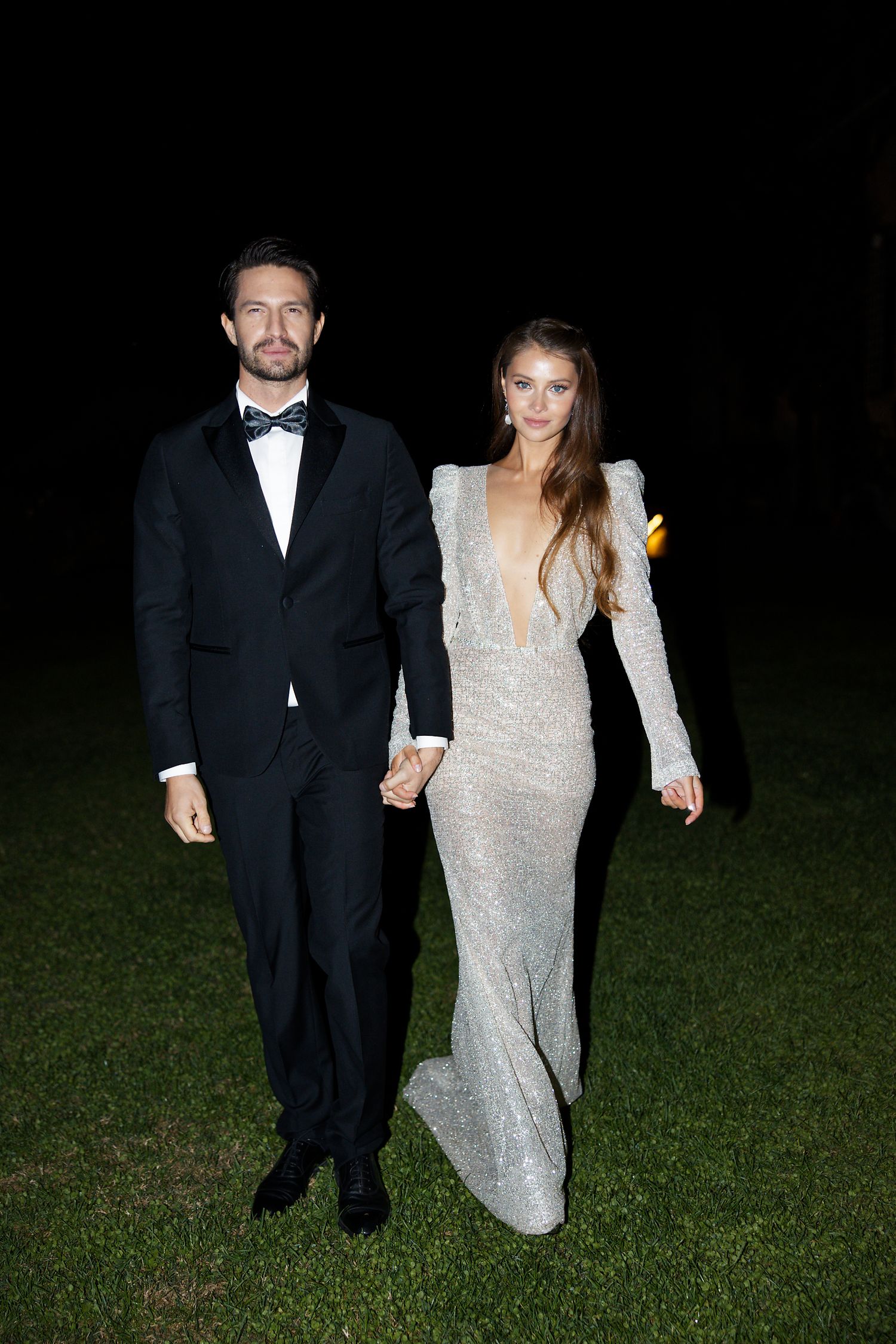 Couple in formal attire holds hands while standing on grass during an evening celebration.