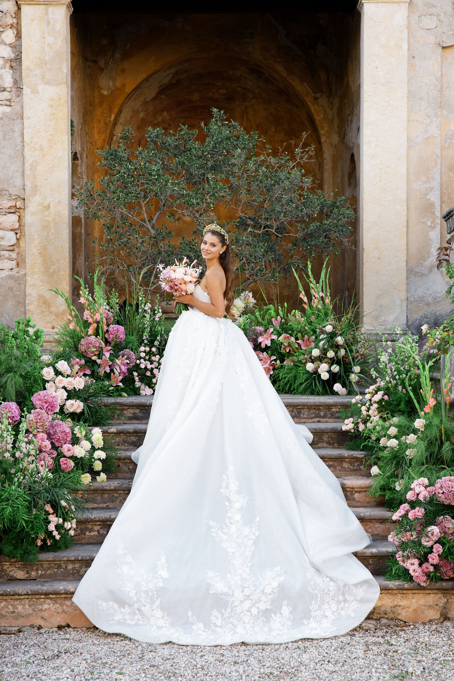 Bride in dramatic white ball gown surrounded by pink flowers and greenery on stone steps.