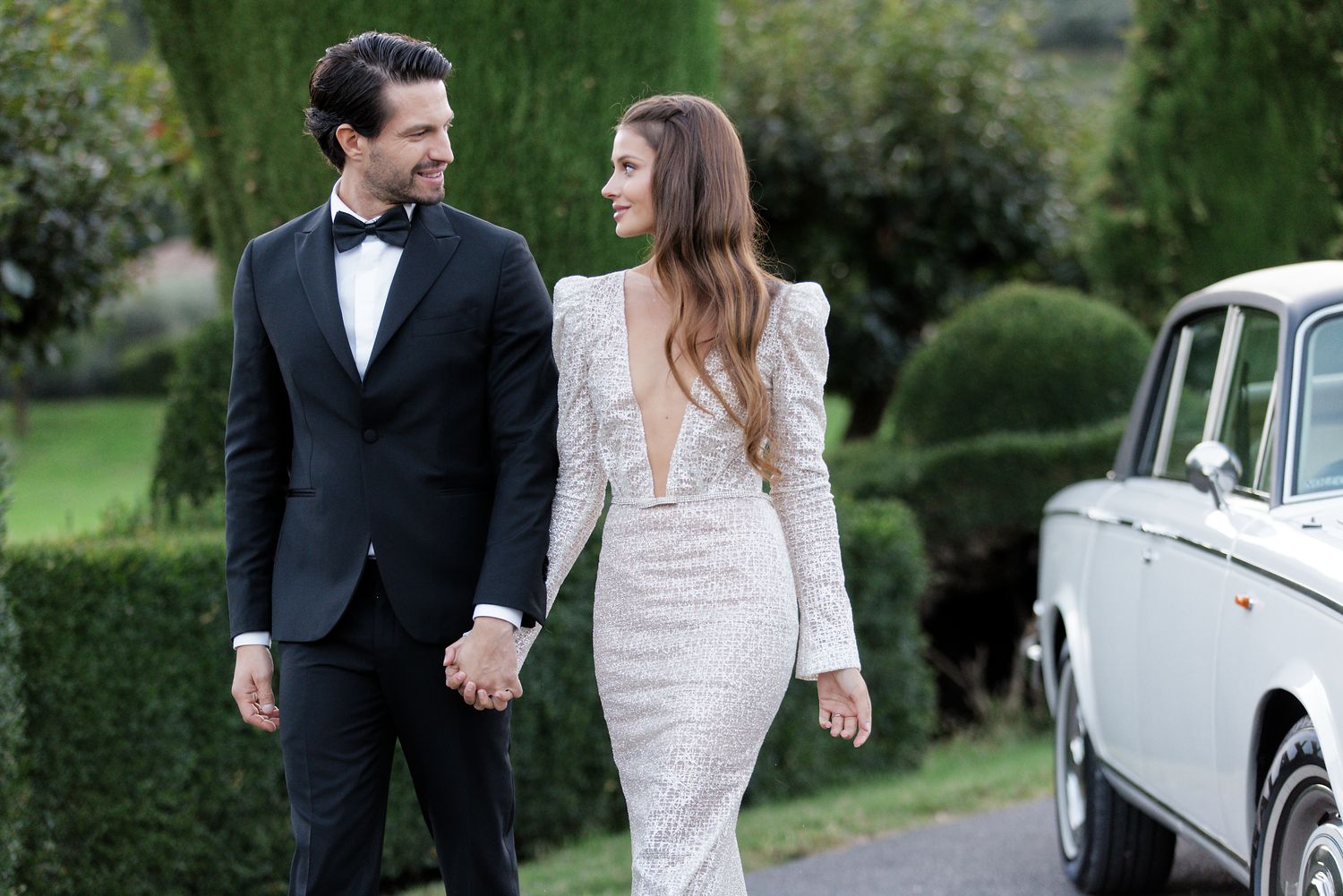 A newlywed couple walks together on a garden path alongside a classic white vintage car.