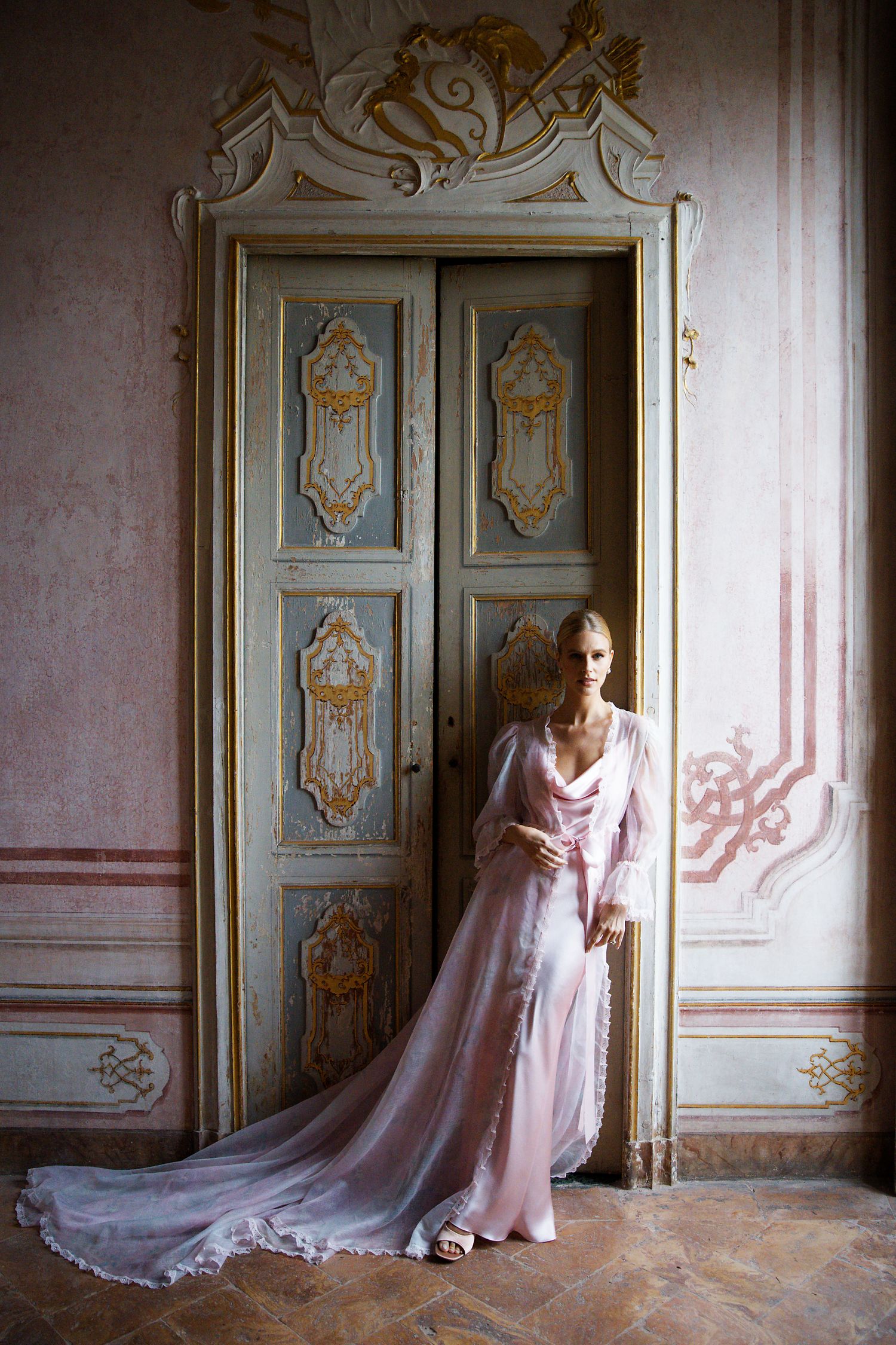 Flowing pink gown with train posed against ornate antique blue doors in historic interior.